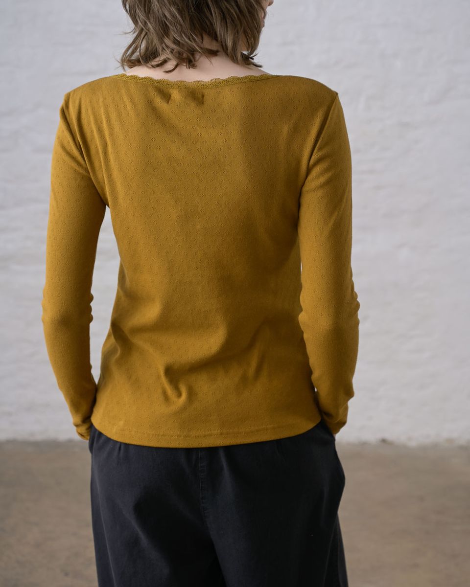 Back view of a woman wearing the BIBICO Cali Pointelle Tee in Bronze, showing the scooped back neckline with scalloped lace trim, fitted silhouette, pointelle eyelet texture, and long sleeves in rich bronze-mustard gold organic cotton, styled with black wide-leg pants against a whitewashed brick wall