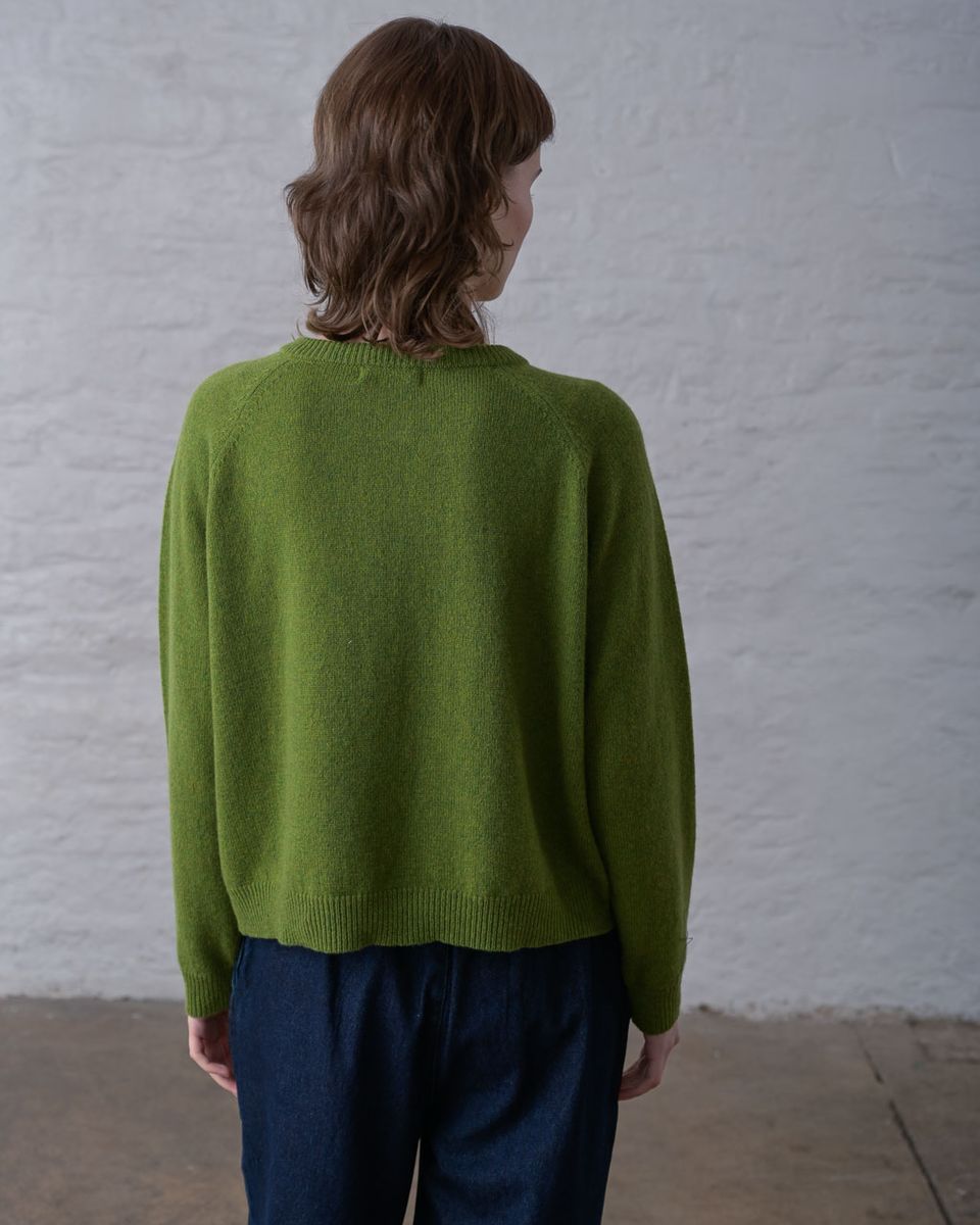 Back view of a woman wearing the BIBICO Lottie Cardigan in Moss, showing the smooth fine knit body, ribbed round neckline, raglan sleeve seams, ribbed cuffs, and straight ribbed hem in midweight moss green lambswool, styled with dark navy wide-leg trousers against a whitewashed brick wall
