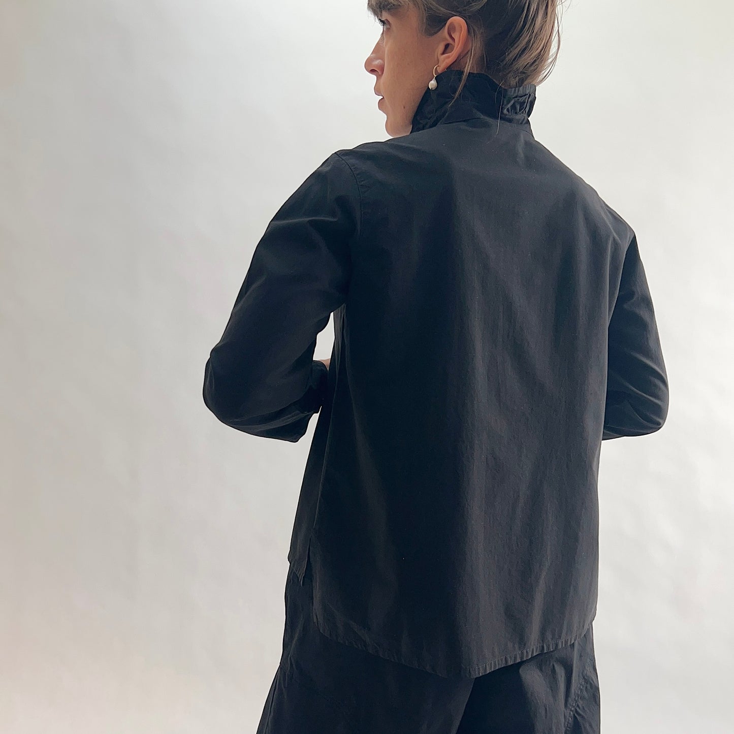 Back view of model wearing Eleven Stitch black blossom collar shirt, showing ruffled collar, long sleeves, relaxed boxy silhouette, curved hem, and black color, styled with matching black wide-leg pants