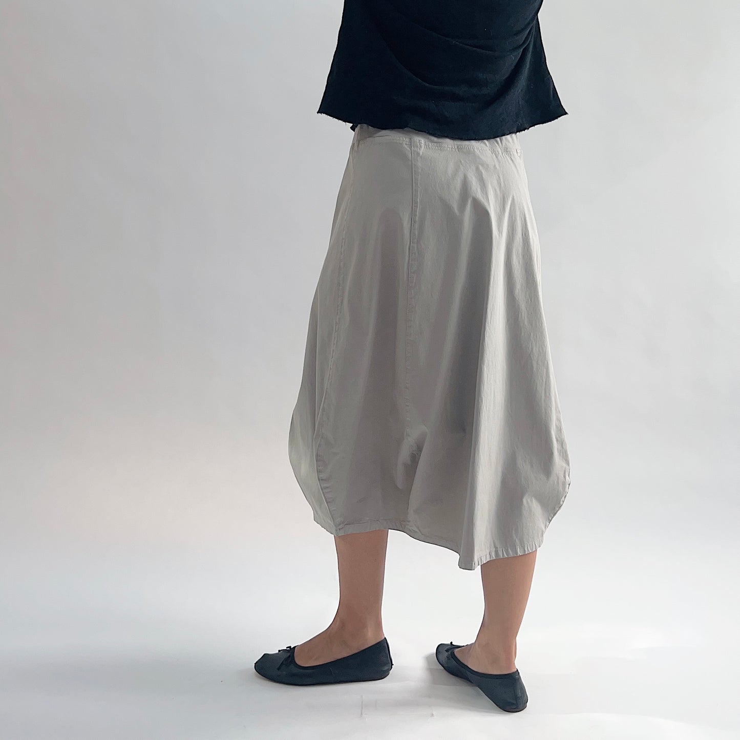 Back view of model wearing Eleven Stitch fennel rib yoke skirt in motion, showing elastic knit waistband, curved seam paneling, sculptural bubble hem, below-the-knee length, and warm taupe color, styled with black top and black ballet flats