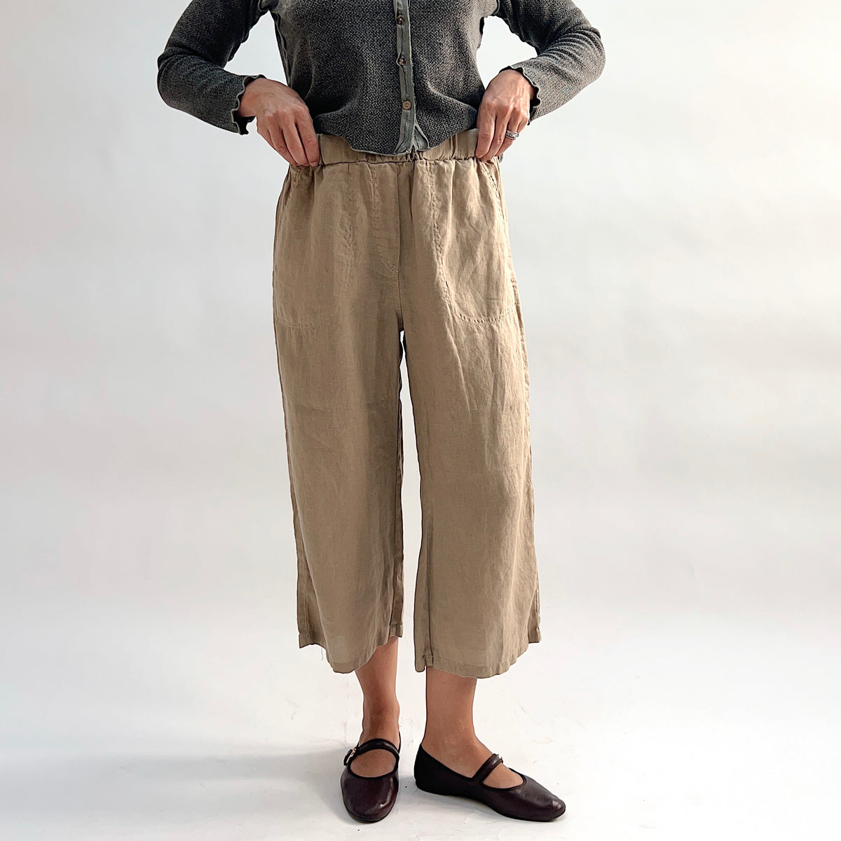 Cut Loose Hanky Linen Easy Crop Flood Pant in Suede, sandy tan linen wide-leg crop pant with hands pulling at elastic waistband, styled with a grey button-front cardigan — front view showing patch pocket and straight hem