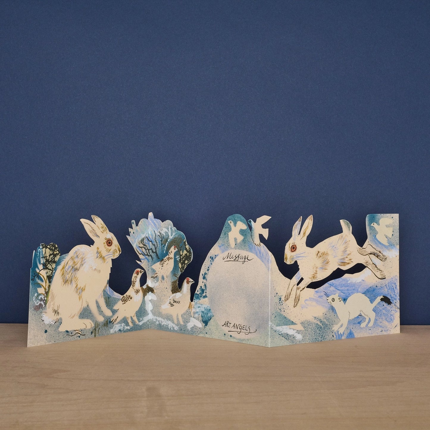 Three Fold Winter Wildlife concertina card open to reveal blank message panel, with a seated white hare and ptarmigan birds on one side and a leaping hare with a stoat on the other, signed by Art Angels