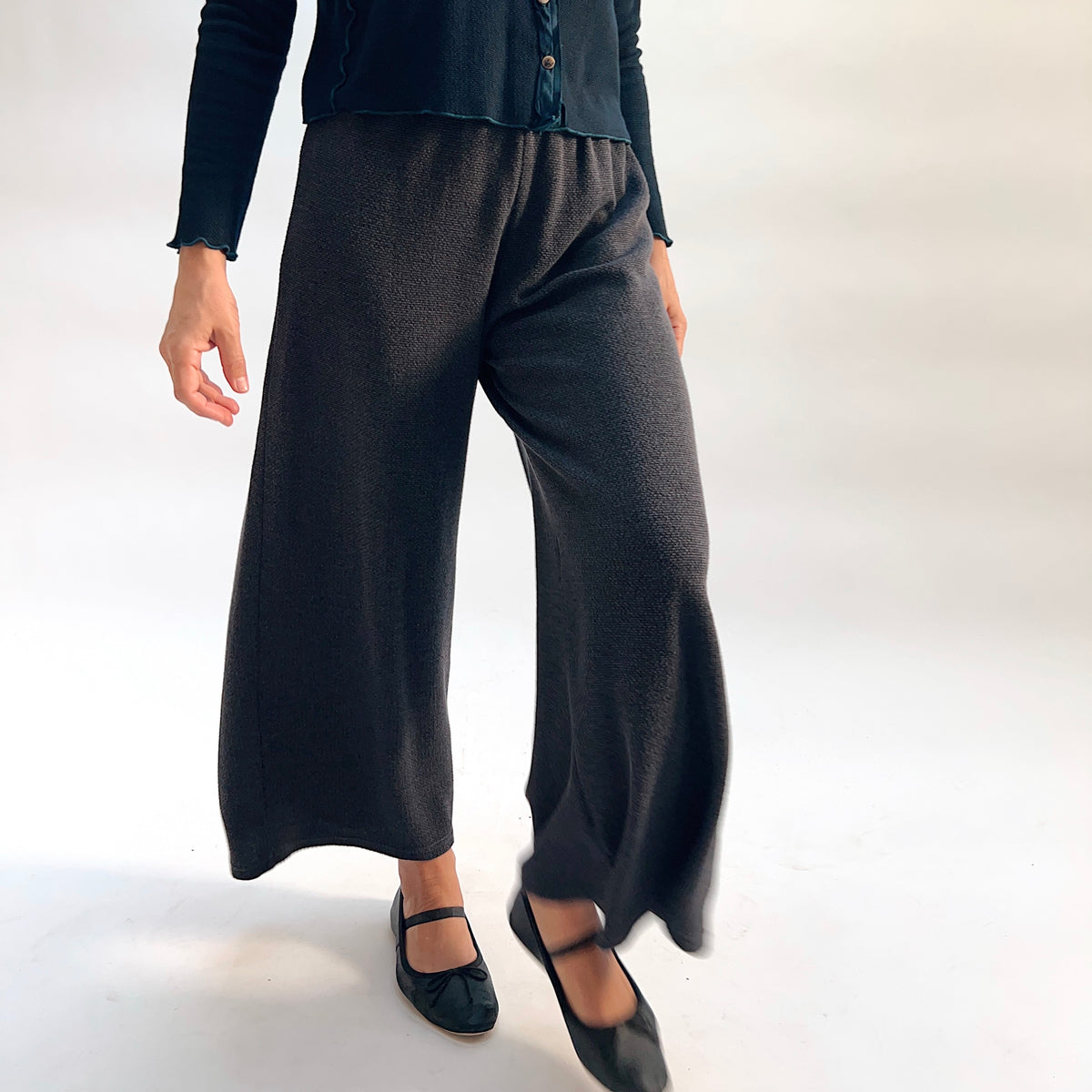 Model wearing the Cut Loose Marled Knit Crop Pant in Iron, close-up side view mid-stride, showing wide-leg silhouette, clean cropped hem, and dark charcoal marled knit texture, styled with a dark teal cardigan and black mary jane flats