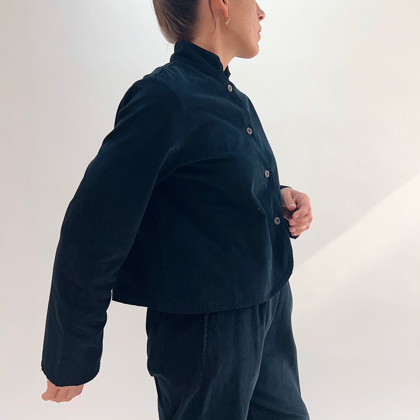 Side profile of model wearing the Bryn Walker Corduroy Standing Collar Crop Jacket in Black, highlighting ruffled standing collar, shell buttons, wide sleeve volume, and cropped silhouette in black pinwheel corduroy, paired with matching pants