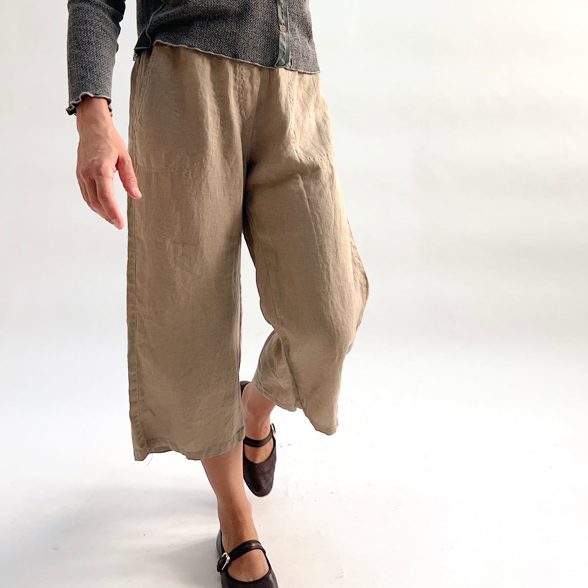 Cut Loose Hanky Linen Easy Crop Flood Pant in Suede, sandy tan linen wide-leg crop pant in motion mid-stride, styled with a grey button-front cardigan and brown Mary Jane flats — front close-up showing linen texture and hem vent