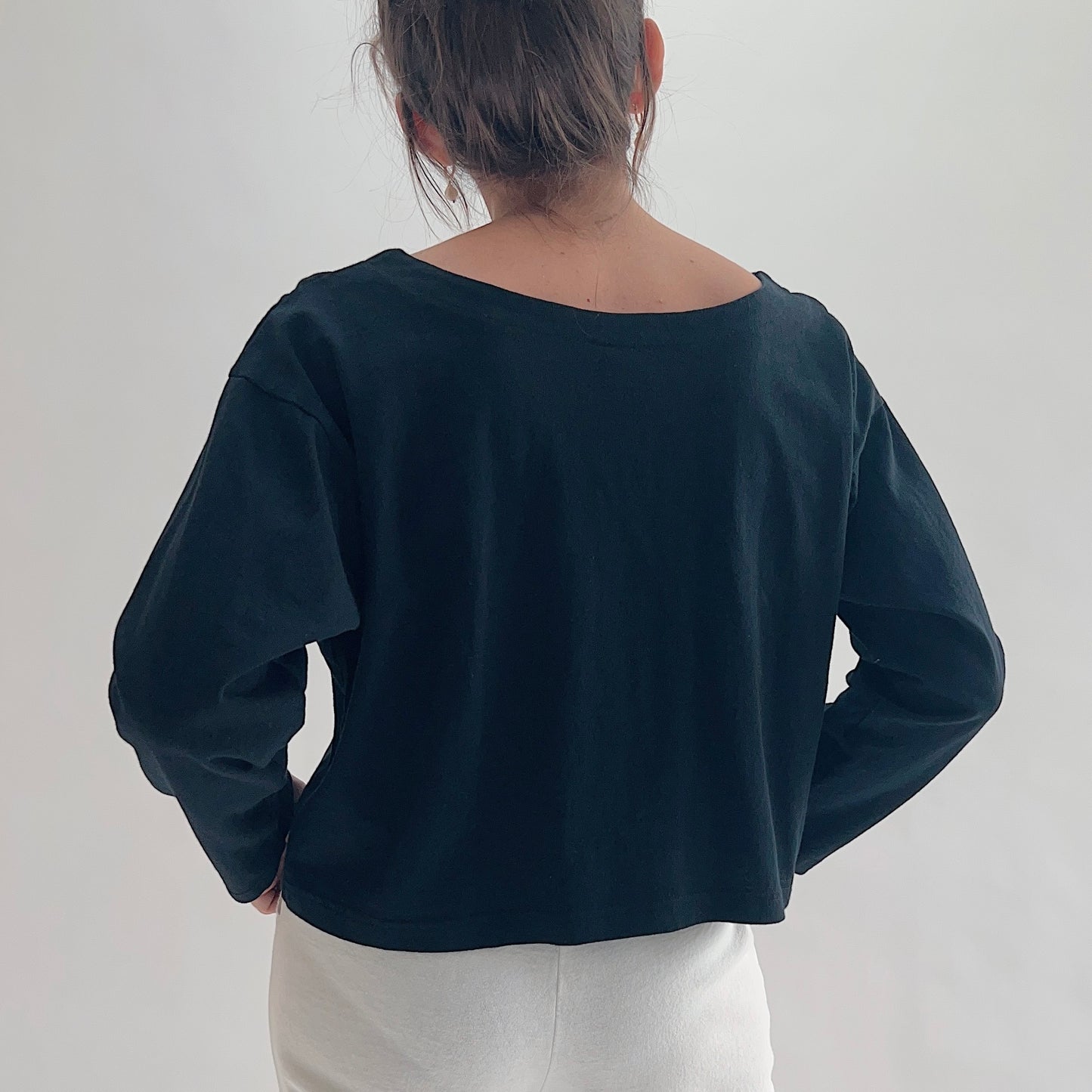 Back view of model wearing Pacific Cotton black long sleeve button front shirt, showing wide scoop back neckline, long sleeves, boxy cropped silhouette, banded hem, and black color