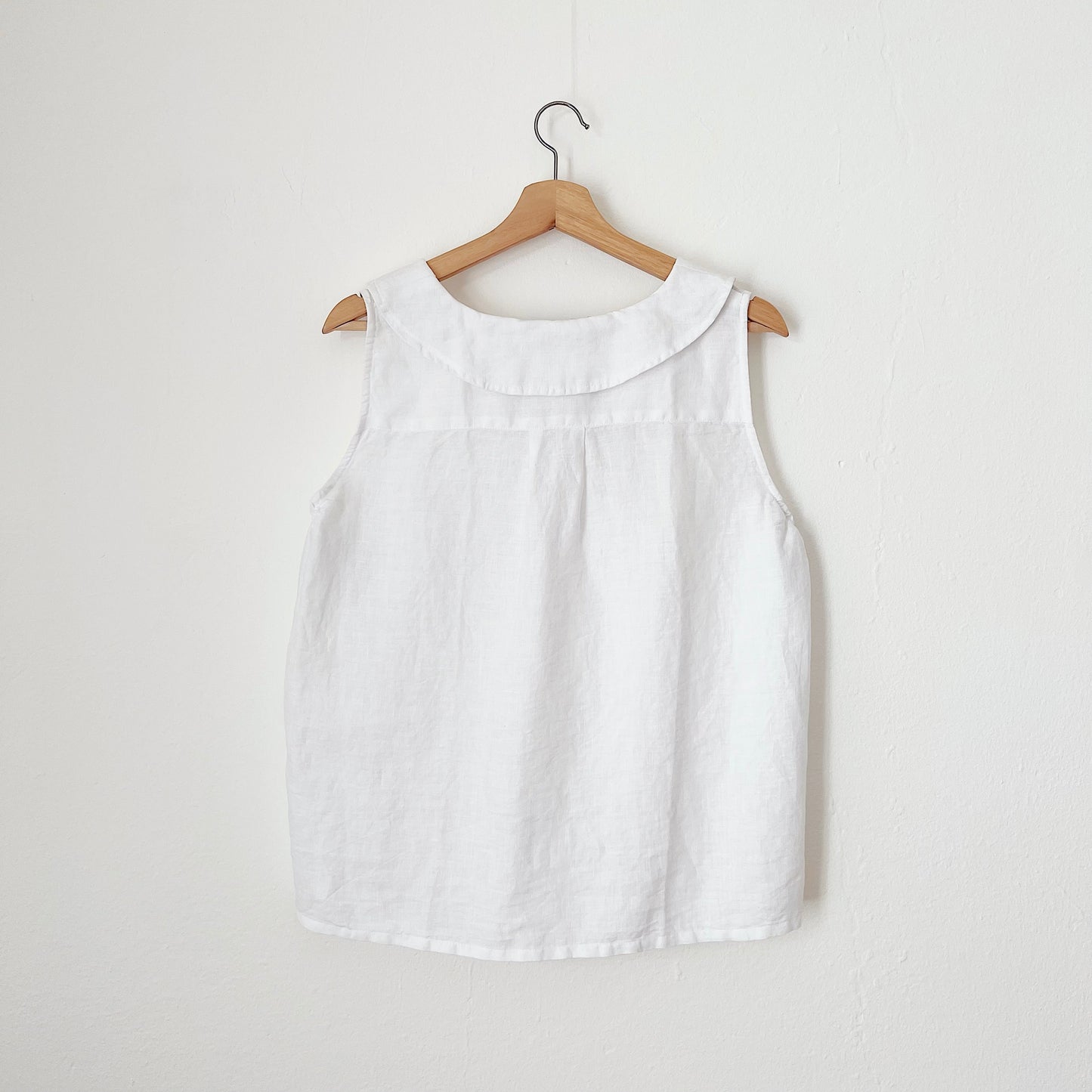 A view of the back of a white linen sleeveless top with a round neckline, hanging on a wooden hanger against a white wall.