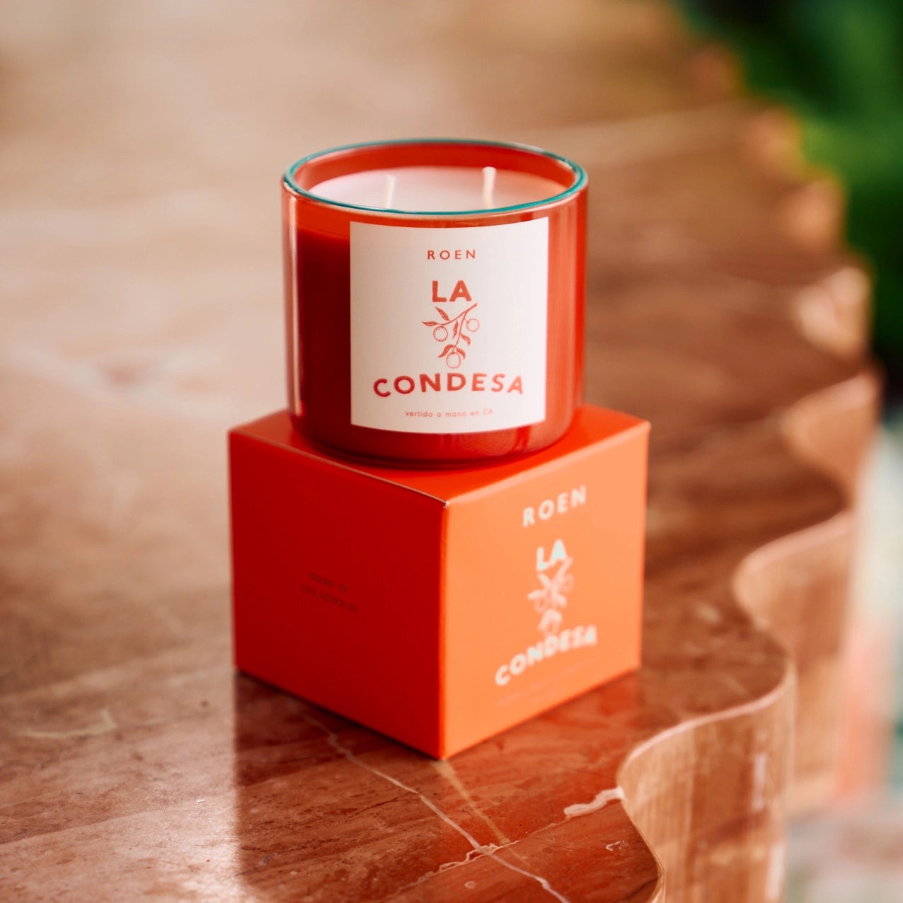 A white candle with a red glass jar, labeled 'ROEN LA CONDESIA', placed on a matching red box, set on a wooden surface.