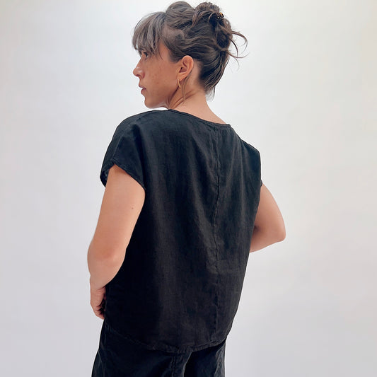 Back view of black linen high-low tee showing center back seam, boatneck, and cap sleeves, hair up with gold hoop earrings, styled with black linen lantern pants