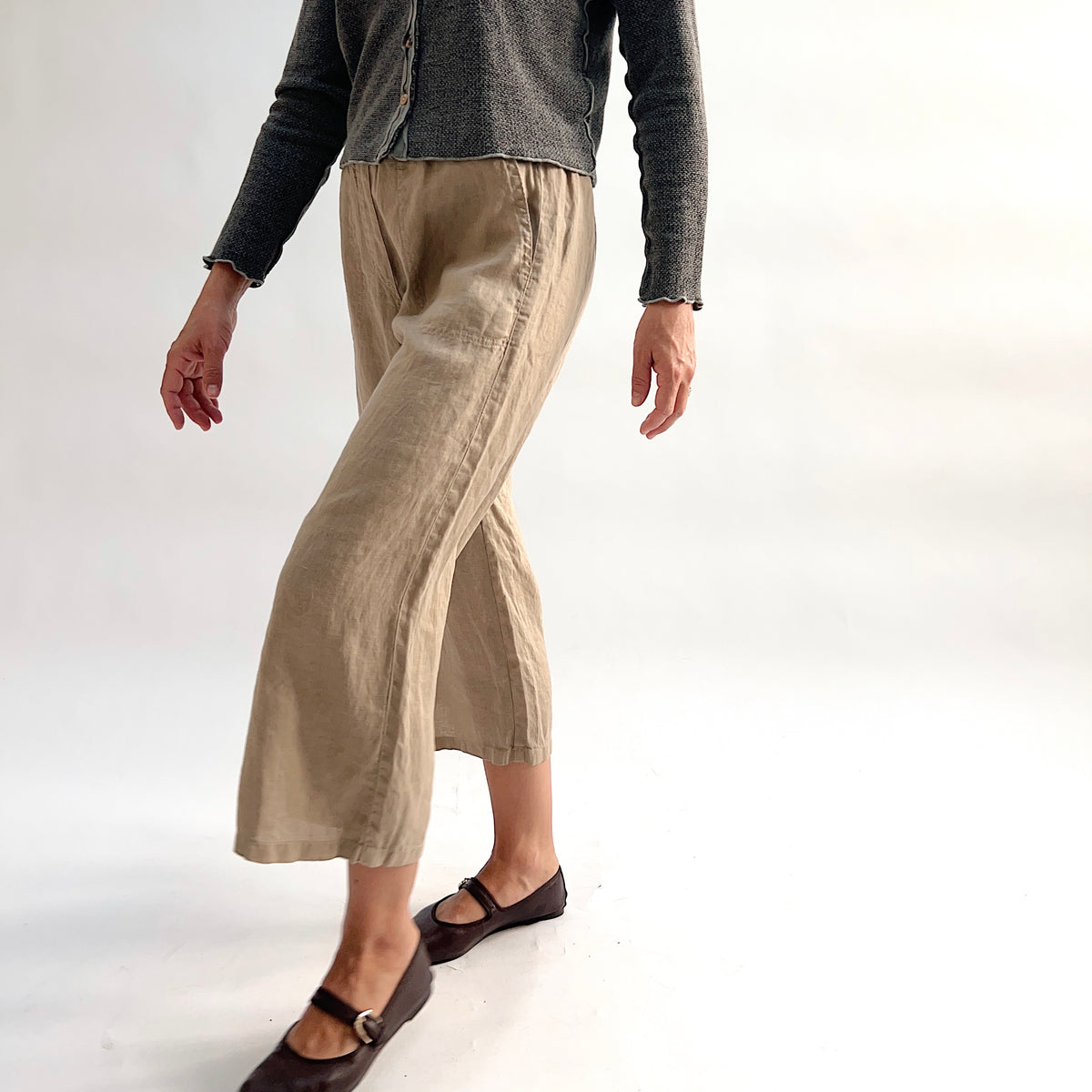 Cut Loose Hanky Linen Easy Crop Flood Pant in Suede, sandy tan 100% linen wide-leg crop pant with front patch pocket, styled with a grey button-front cardigan and brown Mary Jane flats — angled side view showing movement and hem vent