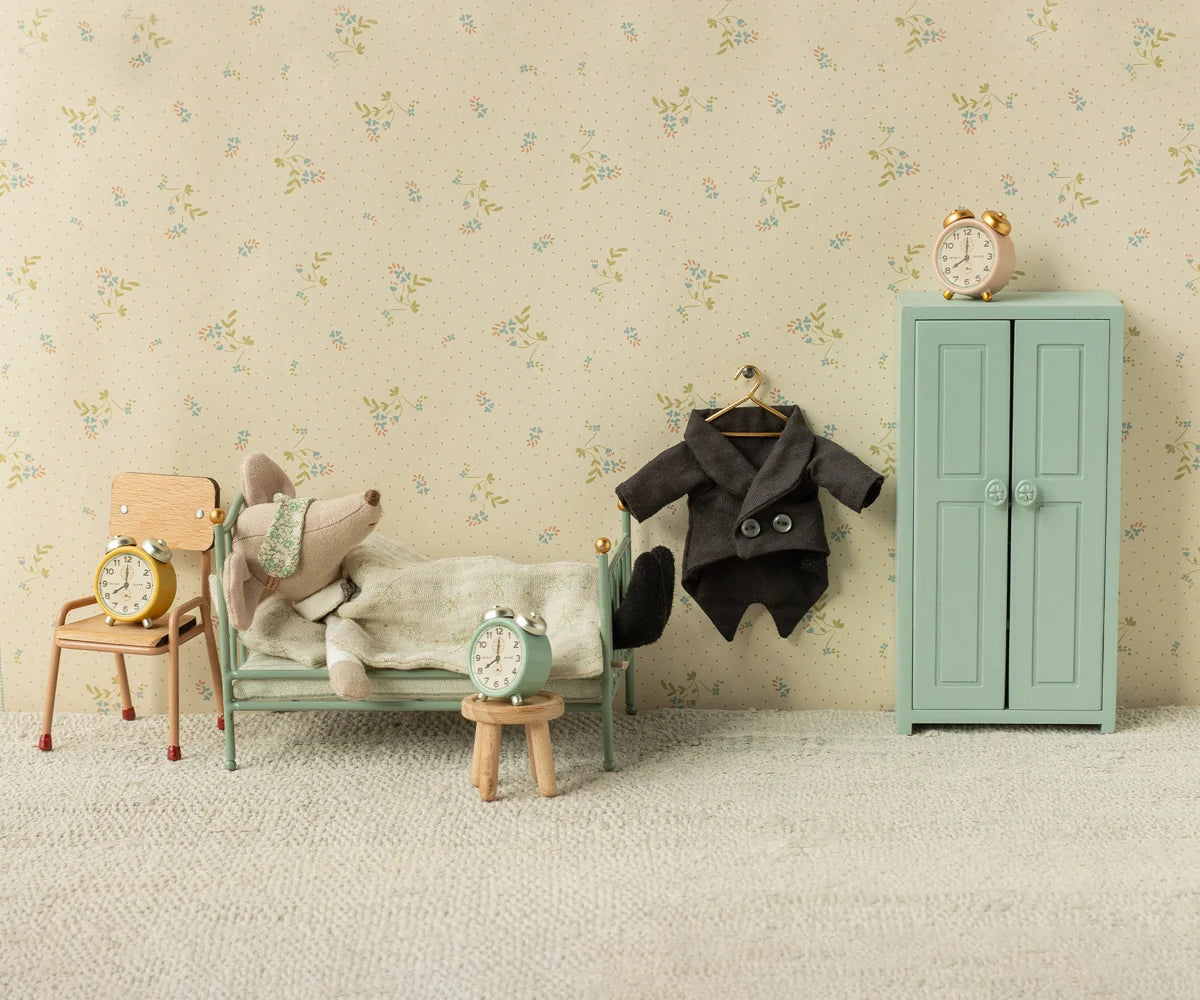 Maileg Vintage Single Bed in Mint — lifestyle scene of a Maileg mouse doll sleeping in the mint bed beside a wooden stool with an alarm clock, a child's chair with a yellow alarm clock, and a teal wardrobe with a dark coat hanging on a hook, against floral wallpaper