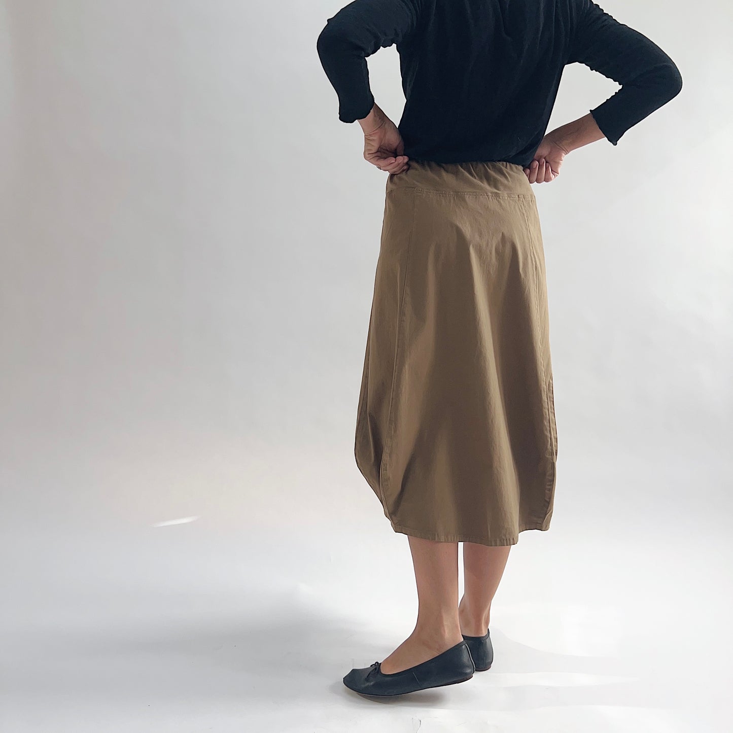Back view of model wearing Eleven Stitch winter olive rib yoke skirt with hands on hips, showing elastic knit waistband, curved seam paneling, sculptural bubble hem, below-the-knee length, and warm khaki olive color, styled with black long sleeve top and black ballet flats
