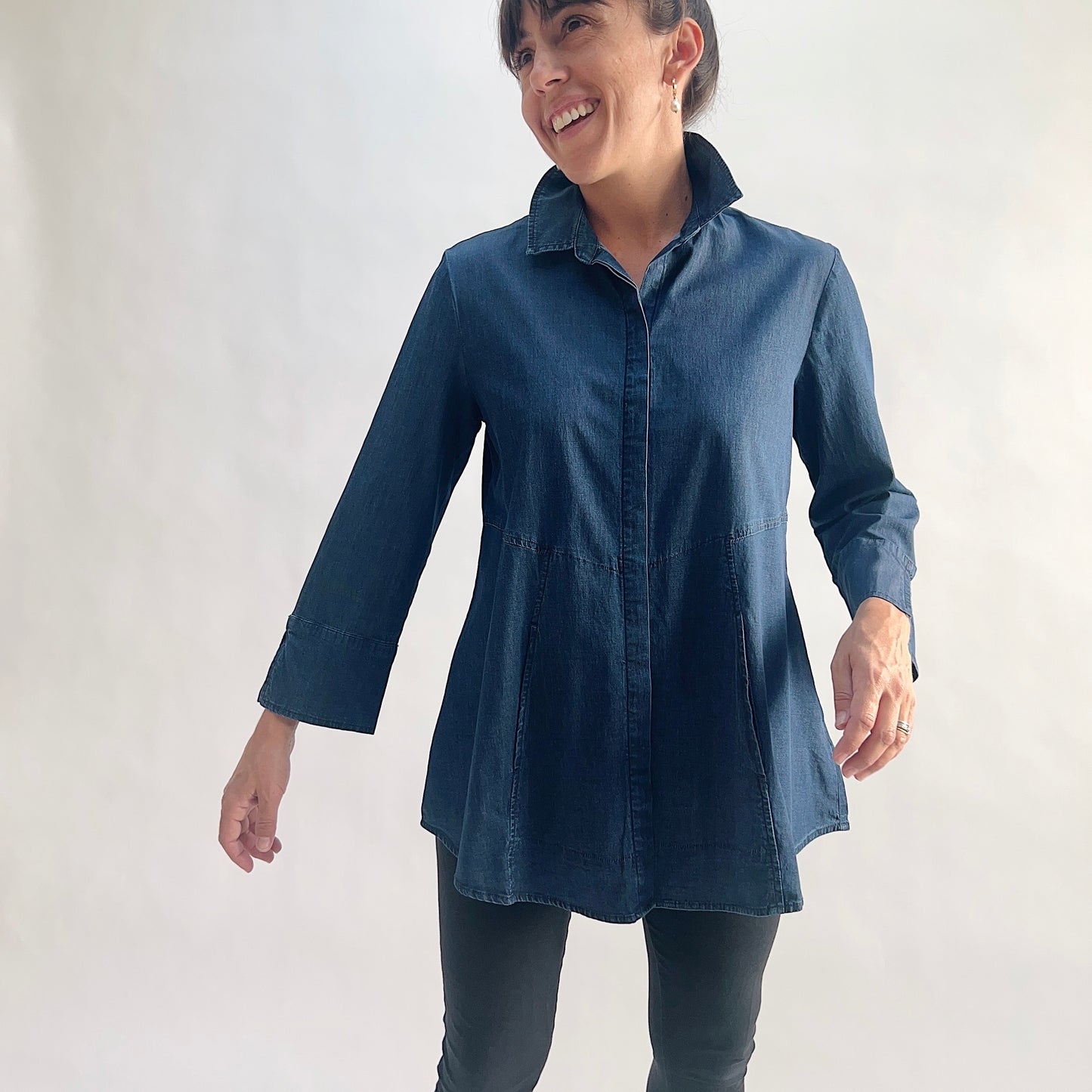 Full front view of model wearing a lightweight dark indigo denim tunic shirt in motion, smiling, showing the hidden front placket, empire seam, flared A-line hem, and 3/4 sleeves with slit cuffs, styled with black slim pants against a white background.
