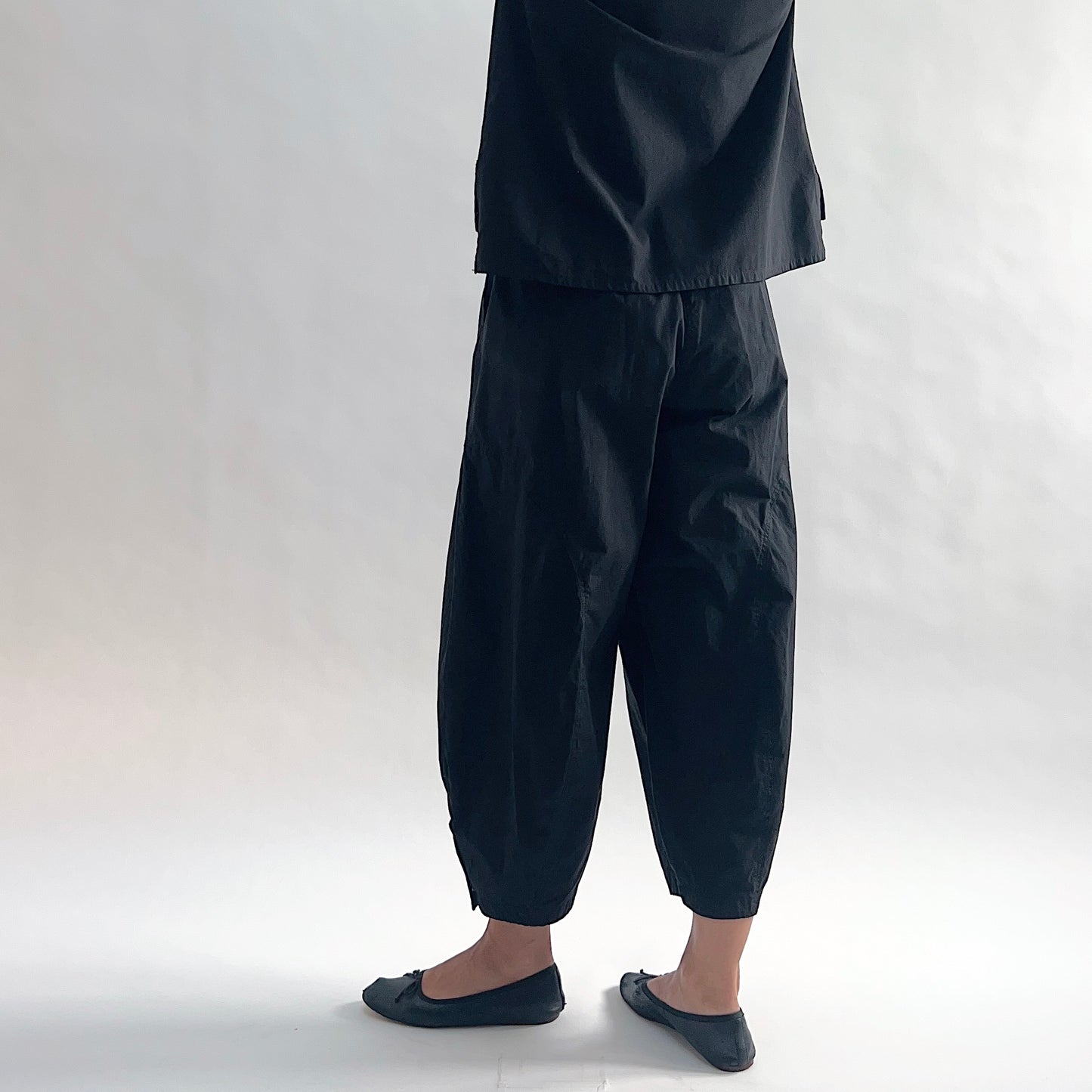 Back view of model wearing Eleven Stitch black snap doreen pant, showing elastic waistband, wide balloon leg tapering to snap hem at ankle, side seam detailing, and black color, styled with matching black top and black ballet flats