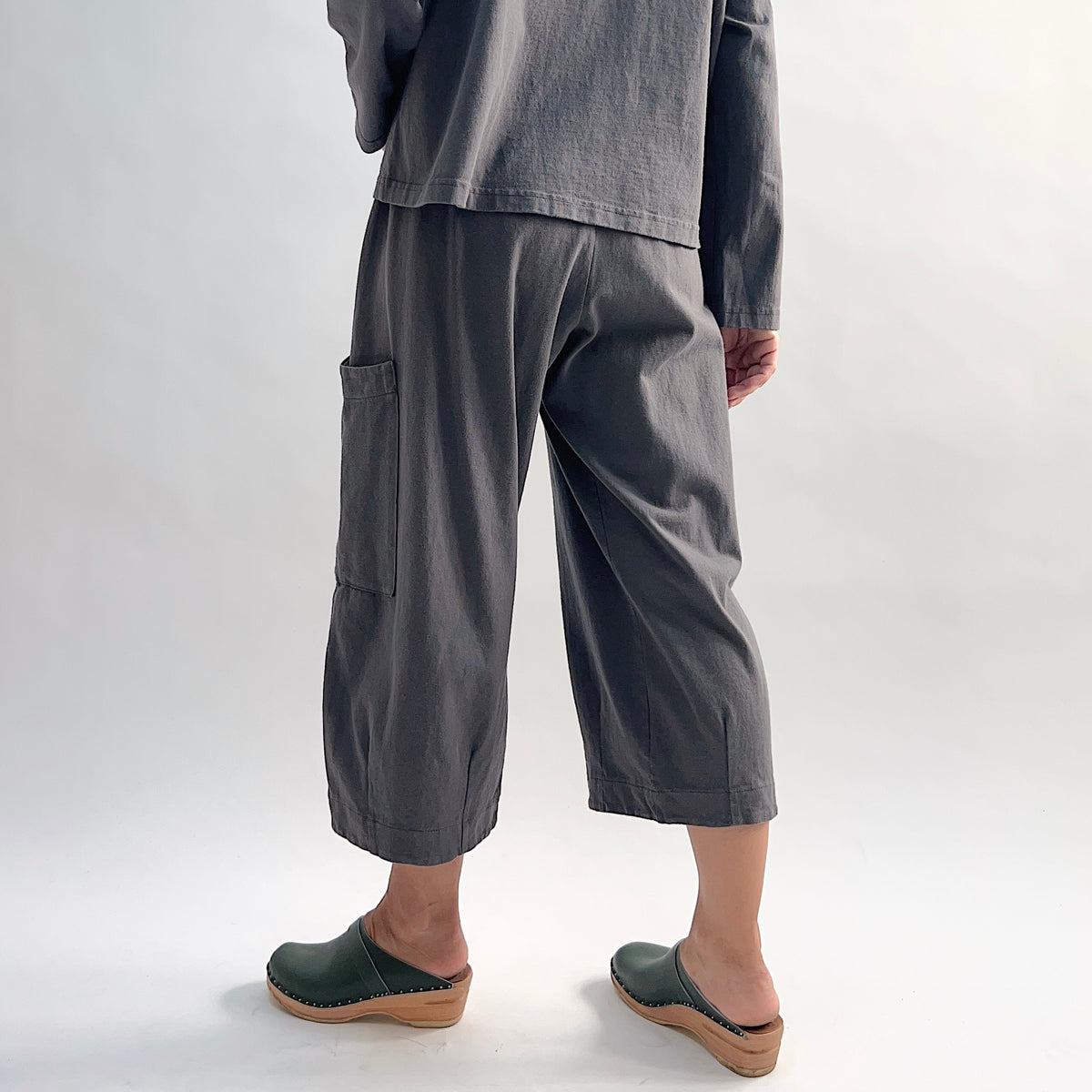 Back view of model wearing the Bryn Walker Cotton Pasha Pant in Moonlight, showing elastic gathered waistband, wide-leg cropped silhouette, thigh patch pocket, folded cuff hem, and soft blue-grey cotton, styled with matching top and green wedge clogs