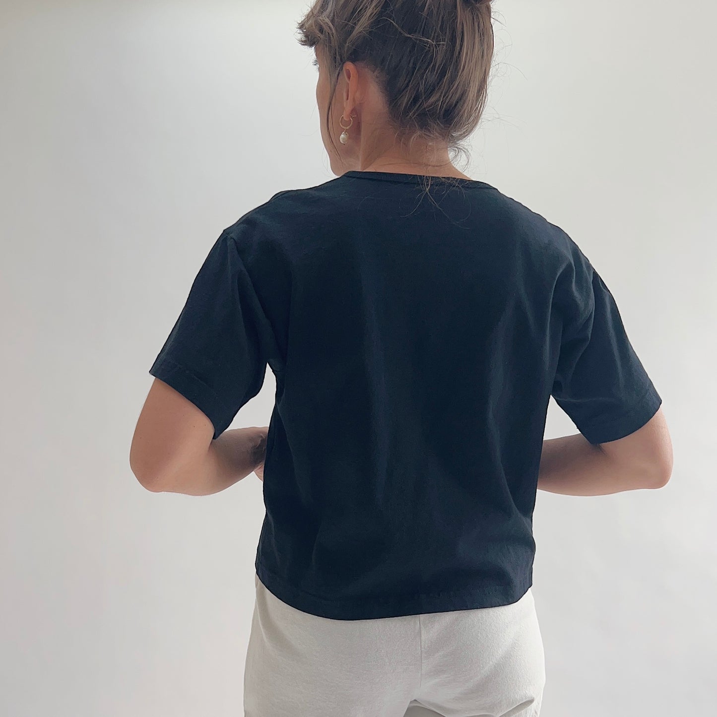 Back view of model wearing Eleven Stitch black crop crew tee with hands behind back, showing crew neckline, short sleeves, relaxed boxy fit, slightly cropped length, and black color, styled with white wide-leg pants