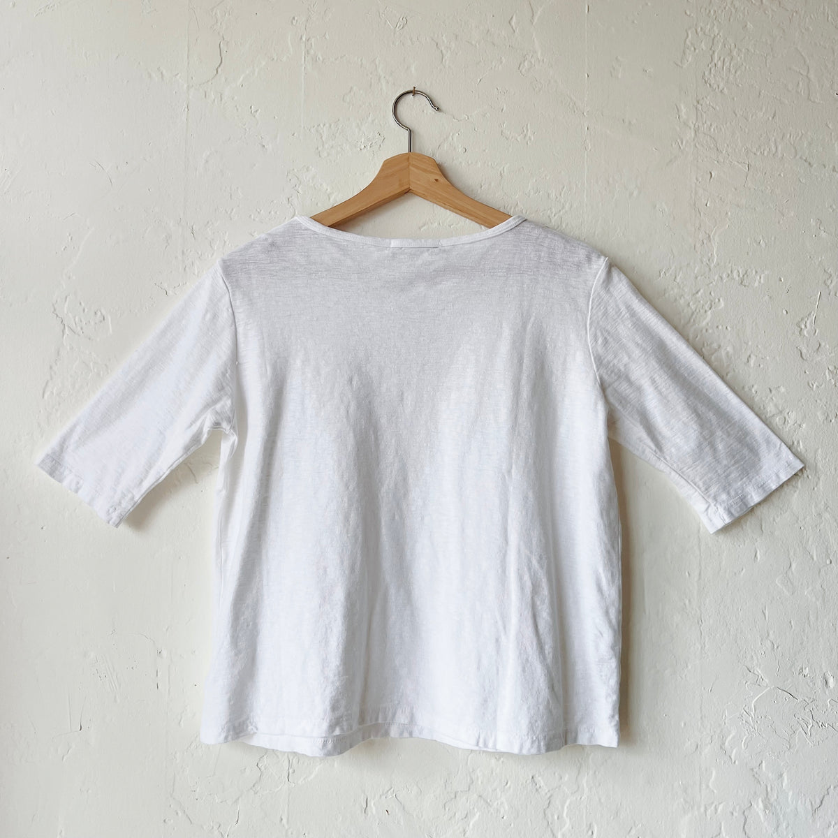 A white elbow sleeve top hanging on a wooden hanger against a textured wall, viewed from the back