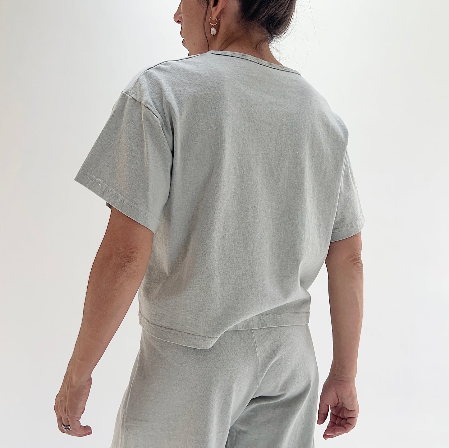 Back view of model wearing Pacific Cotton orlaya crop crew tee, showing short sleeves, boxy cropped silhouette, banded hem, and sage green color