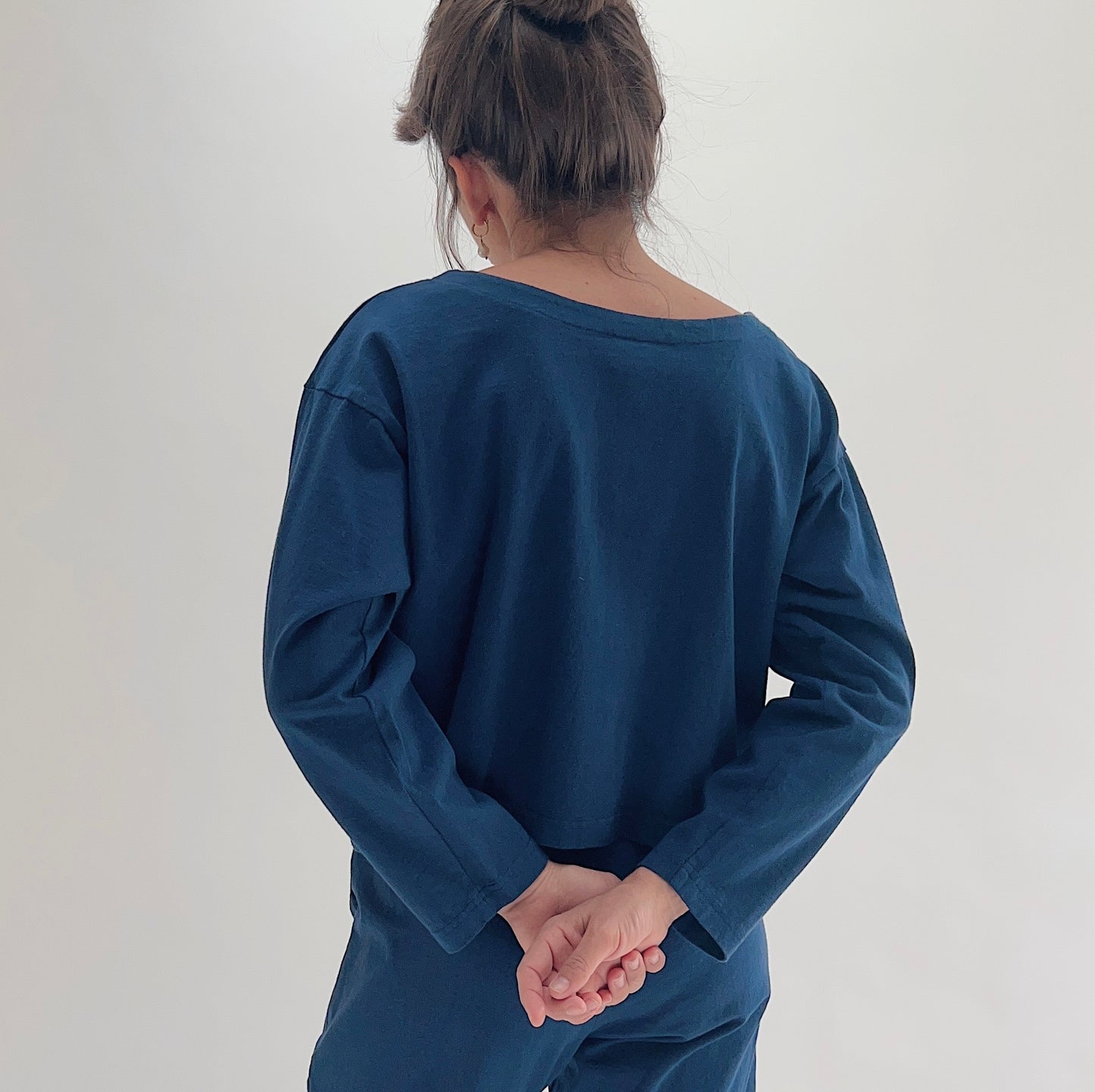 Back view of model wearing Pacific Cotton nova long sleeve button front shirt, showing wide back neckline, long sleeves, boxy cropped silhouette, banded hem, and deep teal color