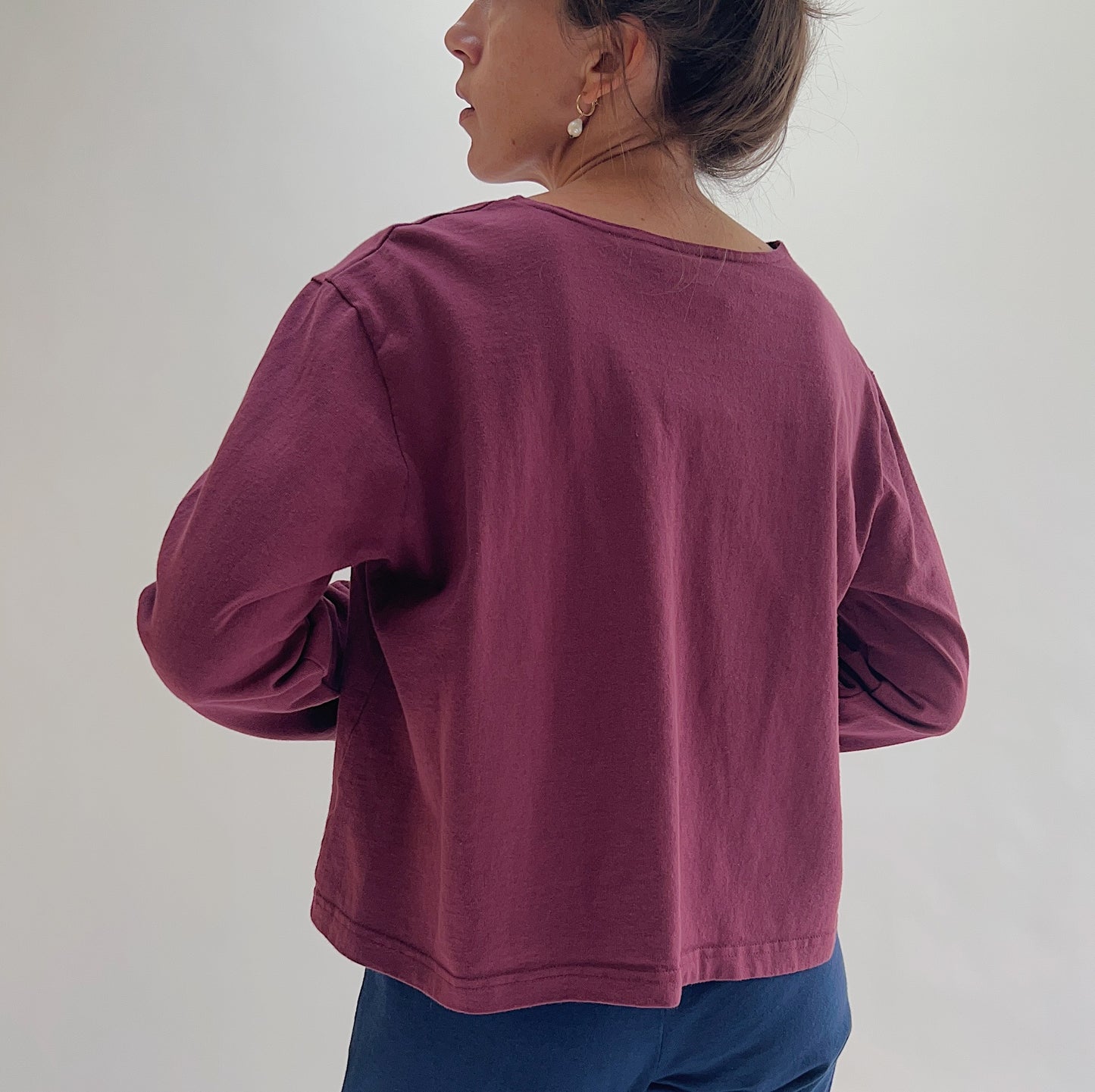 Back view of model wearing Pacific Cotton rhone boxy shirt, showing wide back neckline, long sleeves, boxy cropped silhouette, banded hem, and berry plum color