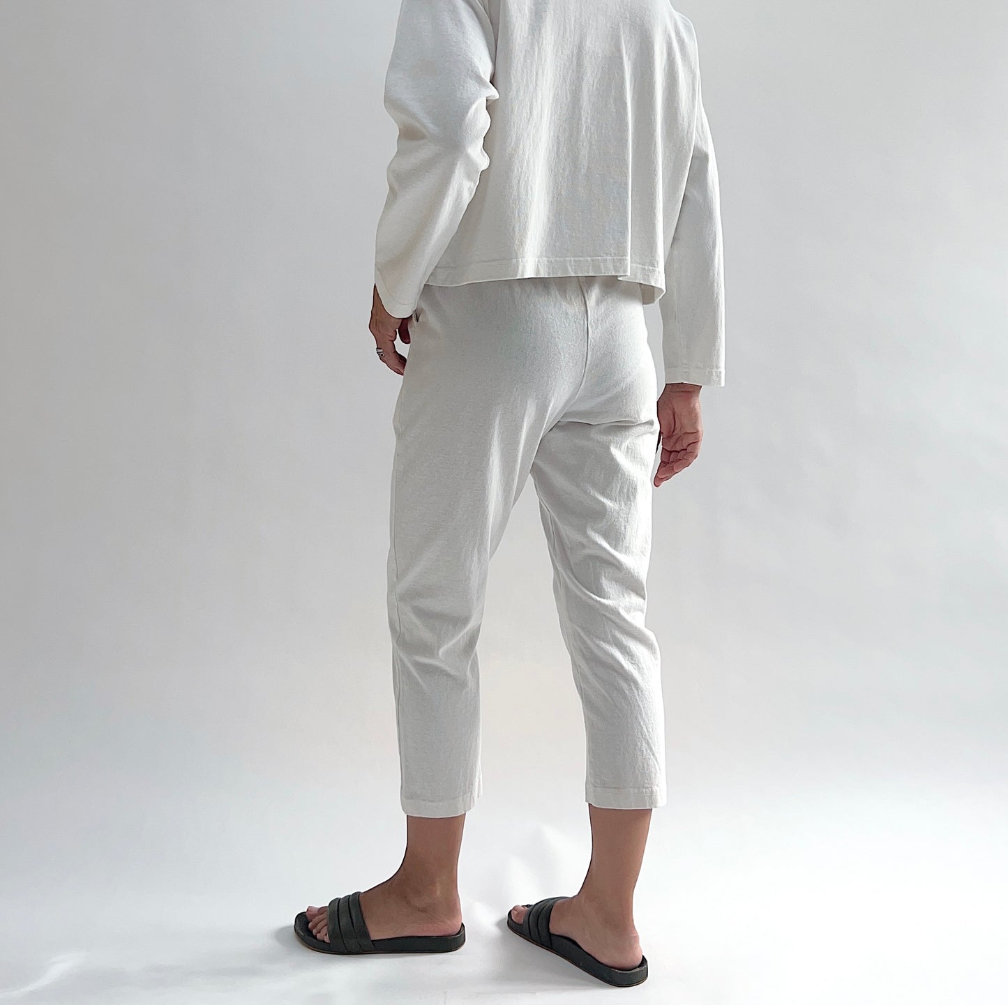 Back view of model wearing Pacific Cotton cream sunday pant, showing elastic waistband, cropped tapered leg silhouette, and off-white color, styled with matching cream long sleeve button front shirt and black slide sandals