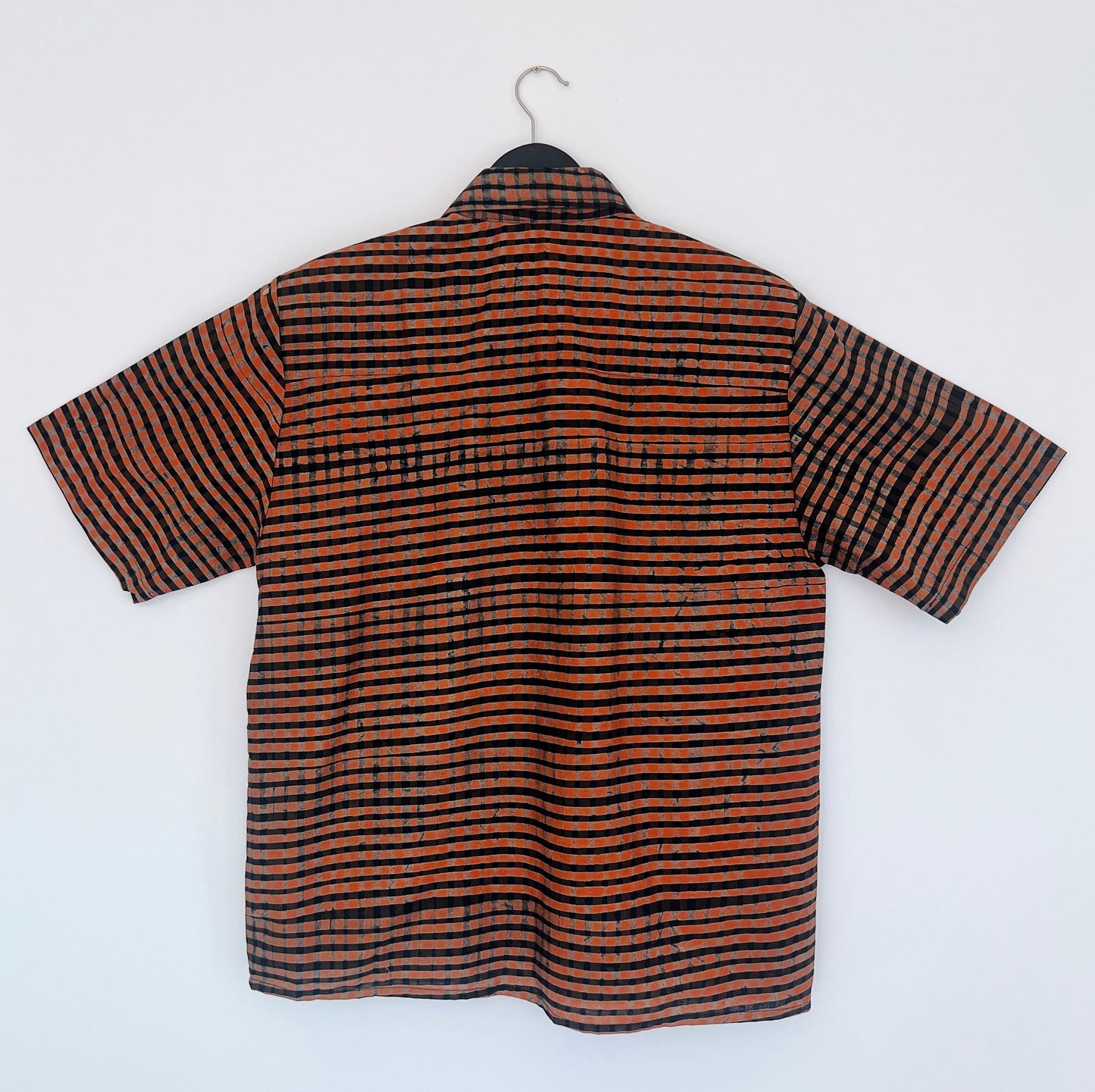 Back view of the Sudi Shirt in Persimmon Grid, hanging on a black hanger against a white wall, showing the relaxed boxy silhouette, short sleeves, and hand-printed persimmon orange and black horizontal grid stripe pattern on 100% cotton Batiki fabric
