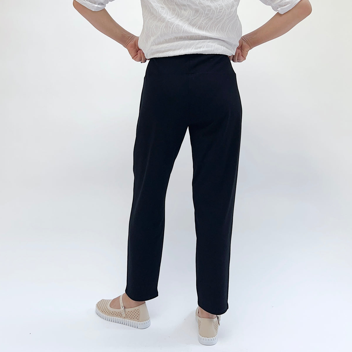 Model wearing the Habitat Core Travel Straight Pant in Black, back view showing contour pull-on waistband, straight ankle leg, and smooth 4-way stretch black knit, styled with white textured top and beige Mary Jane flats