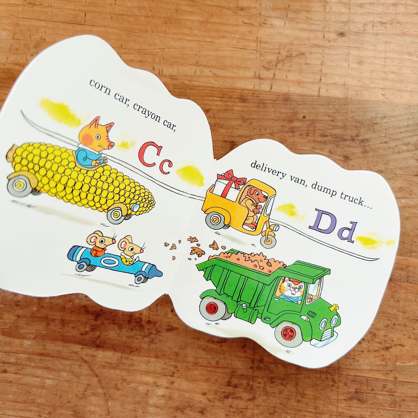 Interior spread of Richard Scarry's Cars and Trucks from A to Z showing illustrated animal characters driving a corn car and crayon car for the letter C, and a yellow delivery van and green dump truck for the letter D