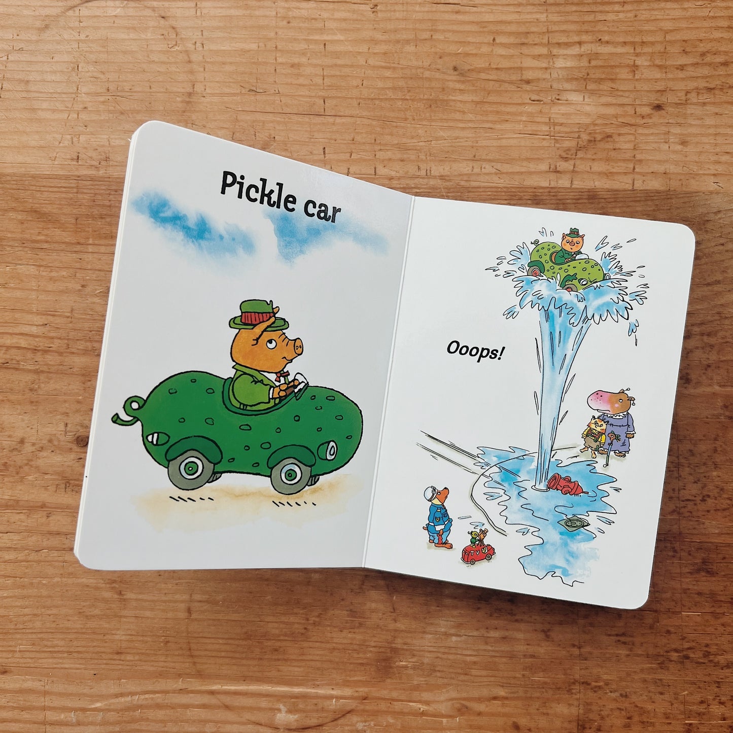 Interior spread of Richard Scarry's Cars board book showing an illustrated pig driving a green pickle car on the left page, labeled "Pickle car," and a comical water hydrant explosion with animal bystanders on the right page, captioned "Ooops!"