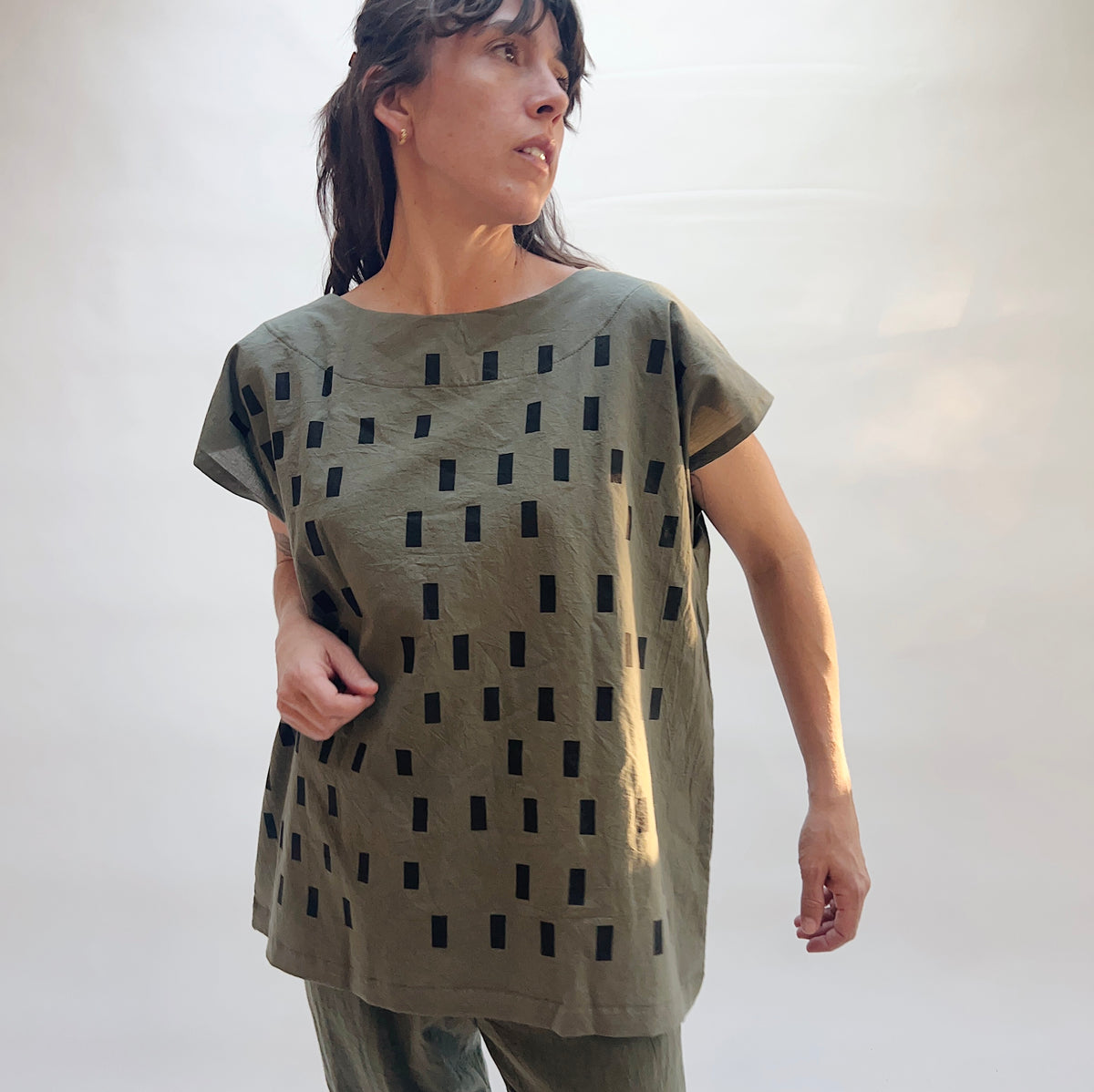Model wearing the Uzi Disko Tunic in Army Green, three-quarter front view, loose army green cotton tunic with allover hand screen-printed black rectangle pattern, capped sleeves, and boat neckline, styled with matching army green pants