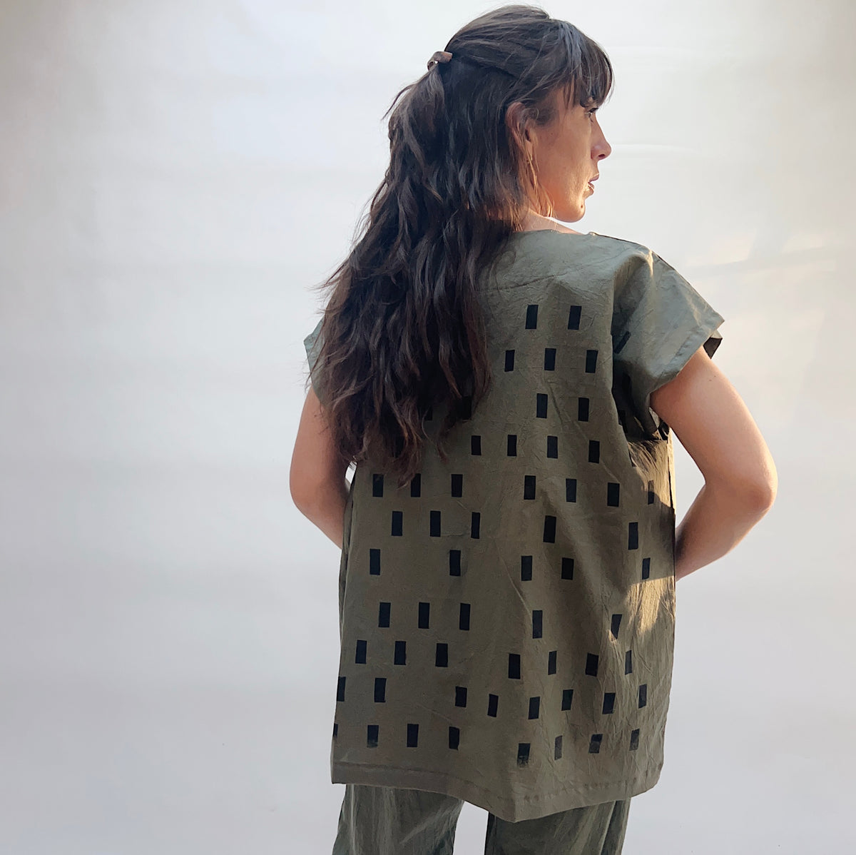 Model wearing the Uzi Disko Tunic in Army Green, back view with hands behind back, showing full hand screen-printed black rectangle pattern on army green cotton tunic and relaxed boxy silhouette, styled with matching army green pants