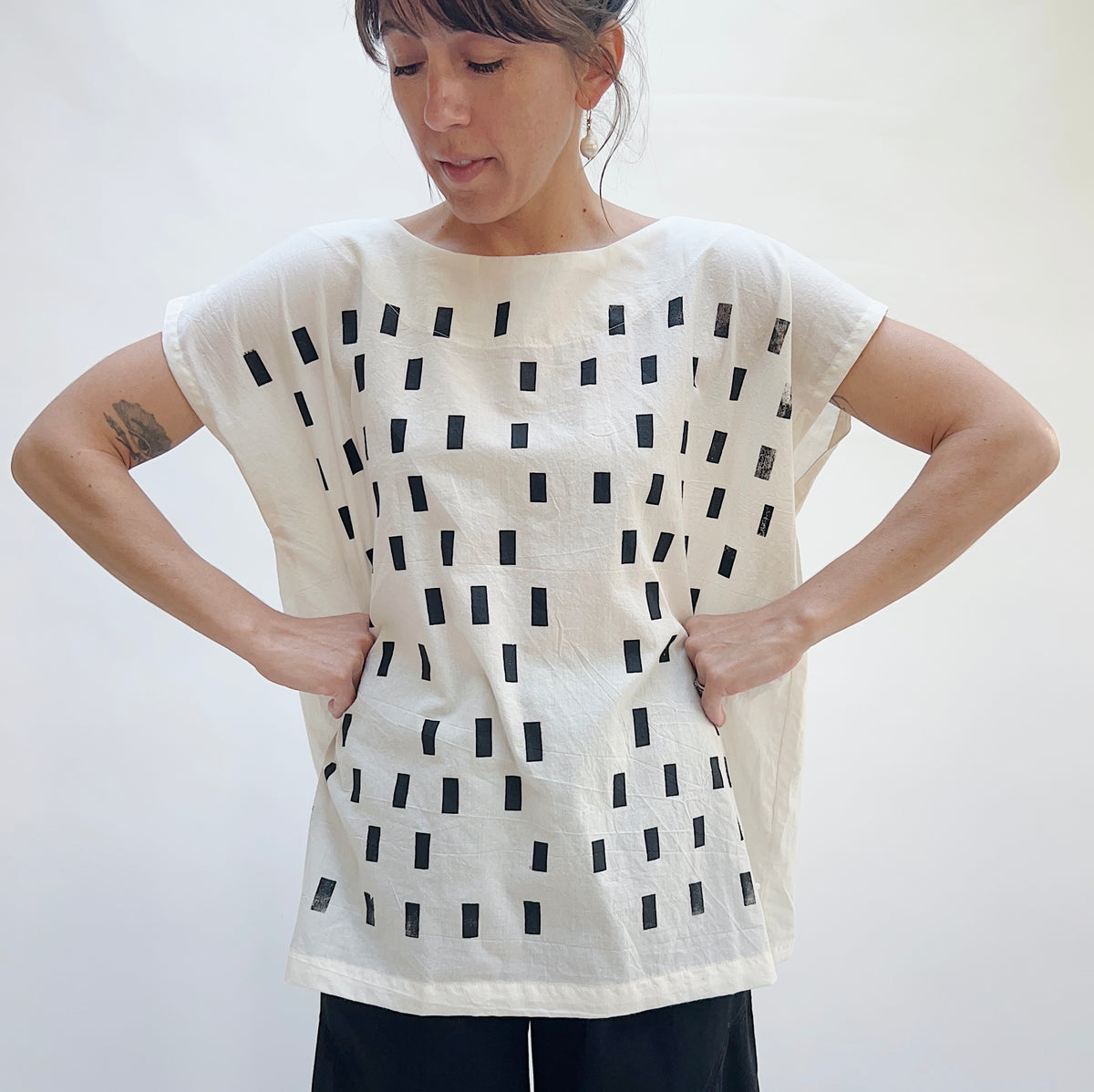 Model wearing the Uzi Disko Tunic in Cream, full-length front view with hands on hips looking down, showing boat neckline, capped sleeves, and allover hand screen-printed black rectangle pattern on cream cotton, styled with black pants