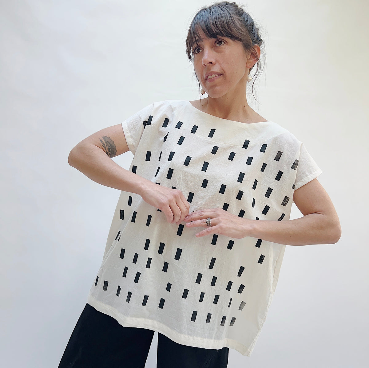 Model wearing the Uzi Disko Tunic in Cream, front view with hands at waist, showing relaxed boxy fit and allover black rectangle screen-print on cream cotton tunic, styled with black pants