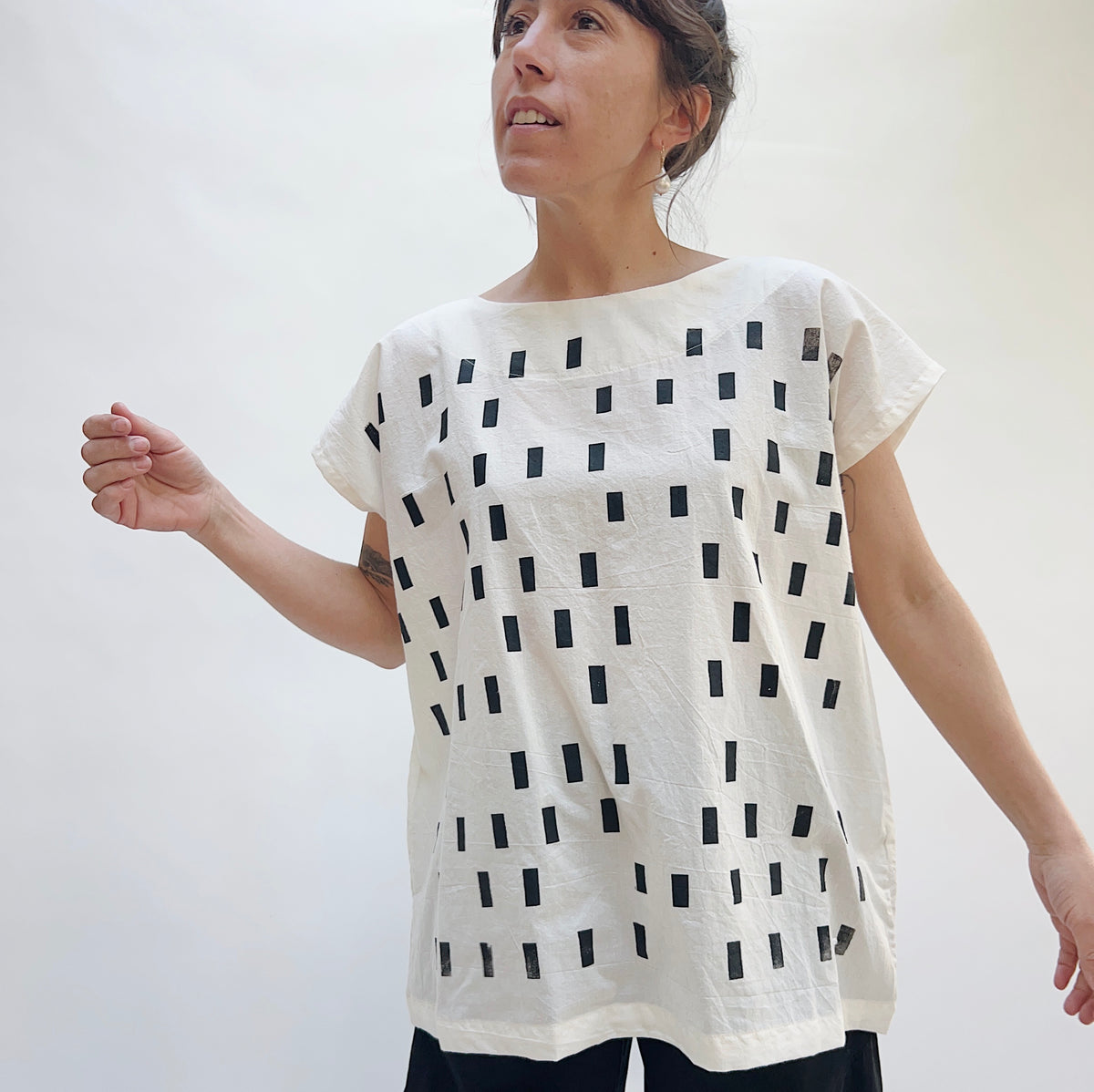 Model wearing the Uzi Disko Tunic in Cream, three-quarter front view looking up, loose cream cotton tunic with allover hand screen-printed black rectangle pattern, boat neckline and capped sleeves, styled with black pants