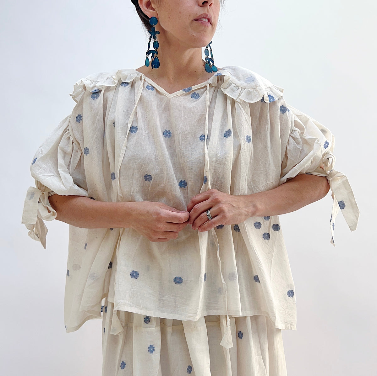 Model wearing DVAA Amelia Top in White Floral, front cropped view against white studio backdrop showing cream 100% cotton with allover woven blue floral spot, oversized scalloped collar with ruffle trim edge and self-tie ribbon at V-neckline, and voluminous elbow-length puff sleeves, styled with matching blue floral spot midi skirt and blue drop earrings