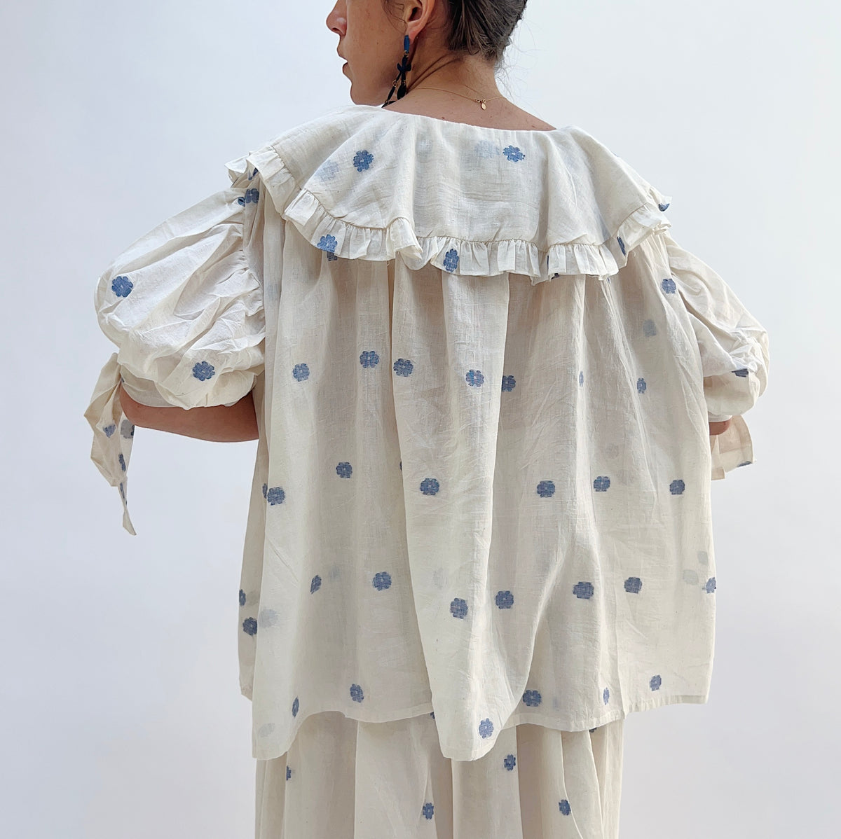 Model wearing DVAA Amelia Top in White Floral, back three-quarter view against white studio backdrop showing cream 100% cotton with allover woven blue floral spot, wide oversized scalloped collar with ruffle trim edge across the upper back, voluminous elbow-length puff sleeves with self-tie ribbon cuffs, and relaxed gathered back, styled with matching blue floral spot midi skirt and gold necklace