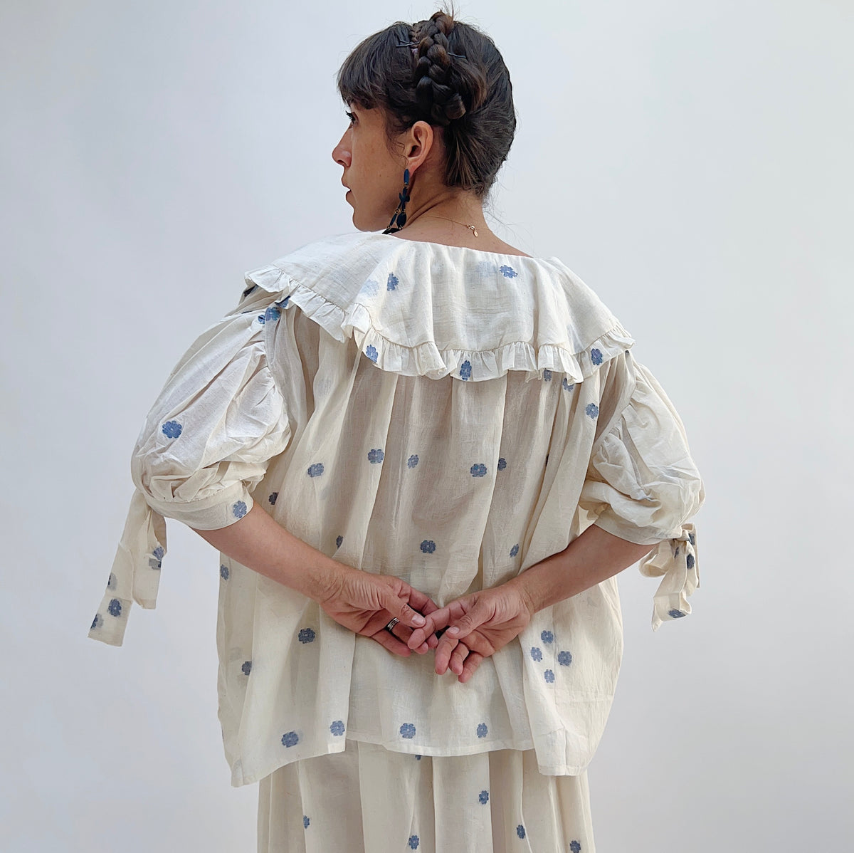 Model wearing DVAA Amelia Top in White Floral, full back view against white studio backdrop showing cream 100% cotton with allover woven blue floral spot, wide oversized scalloped collar with ruffle trim across upper back, voluminous elbow-length puff sleeves with self-tie ribbon cuffs, relaxed gathered boxy back, styled with matching blue floral spot midi skirt and blue drop earrings