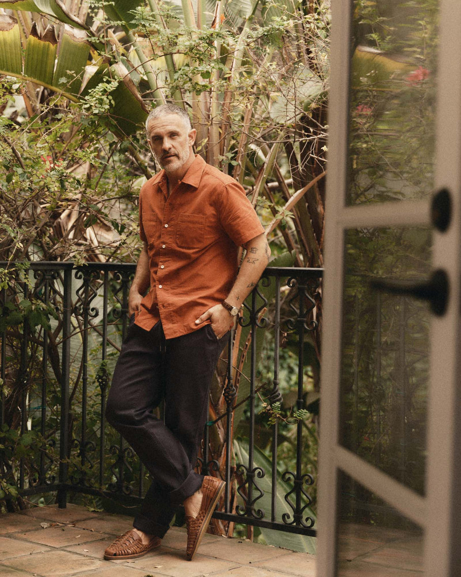Model wearing Taylor Stitch Conrad Shirt in Heather Chili Pincord, full-length view on lush garden balcony showing warm heather chili organic cotton pincord, open camp collar, short sleeves, two chest pockets, and straight hem, styled with dark navy trousers and woven leather huaraches