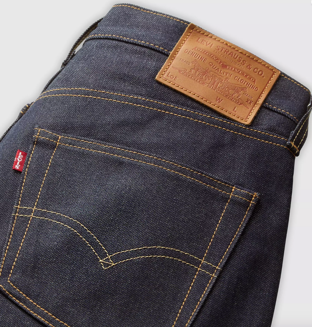 Close-up of the back waistband and pocket area of Levi's dark indigo denim jeans, showing the tan leather patch, red tab, and contrast stitching on a light gray background.