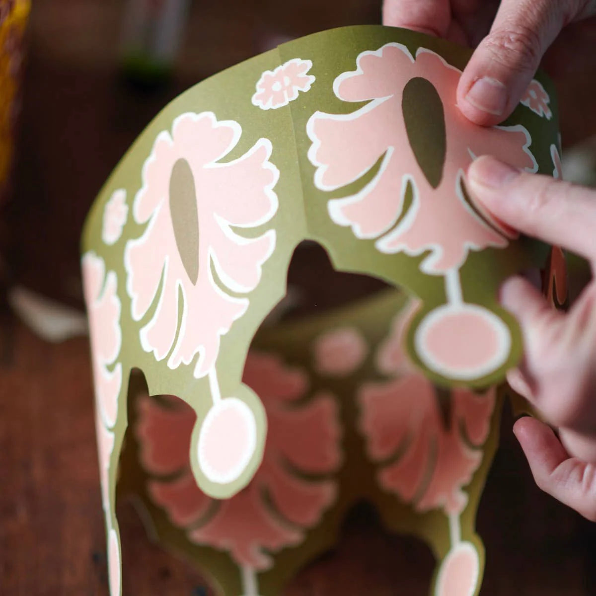 Hand holding a decorative paper cutout of a fish with floral patterns on a wooden surface.