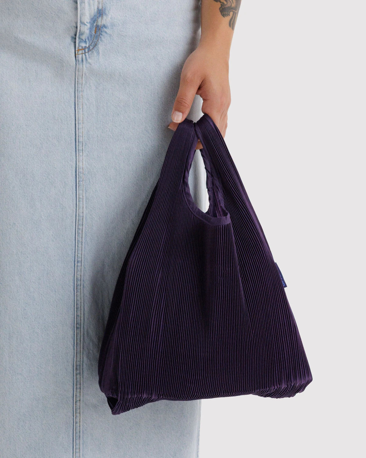 BAGGU Pleated Baggu in Fig, held at a model’s side to show the dark purple bag’s pleated texture and shape.