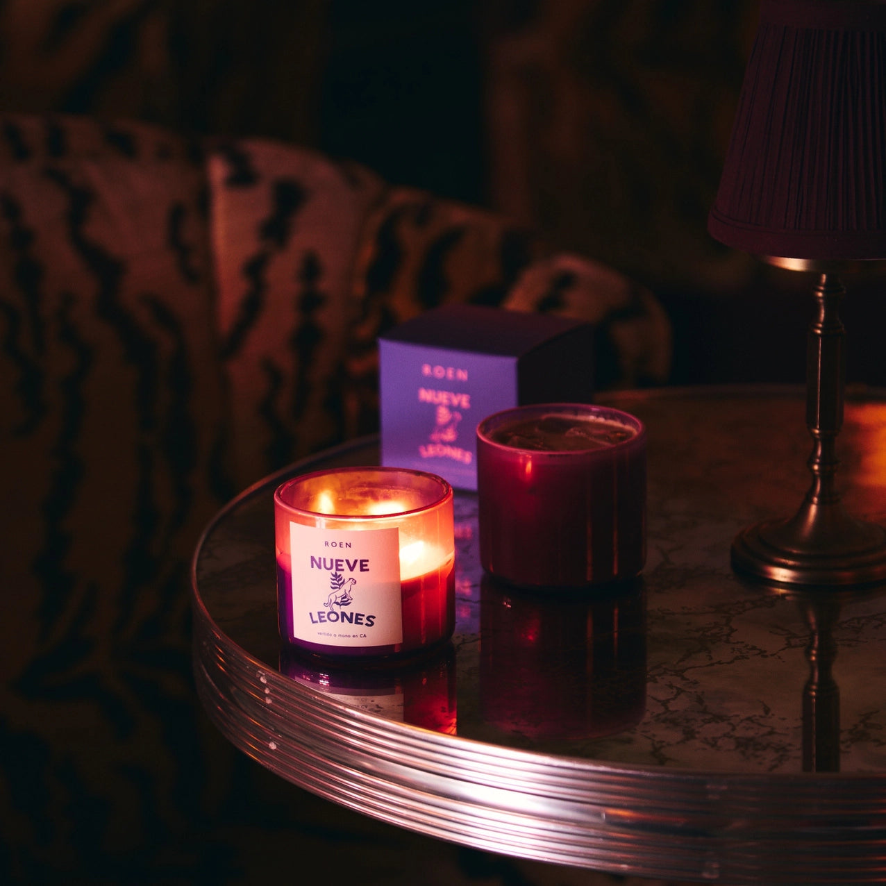 A Roen candle with a label 'NUEVE LEONES' burning on a mirrored table next to a brass lamp