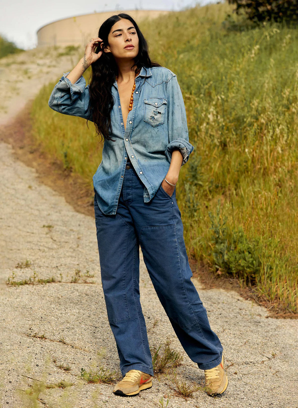 Full-length view of model wearing blue Clarke Canvas Workpants with a chambray shirt and gold sneakers, standing on a gravel road.