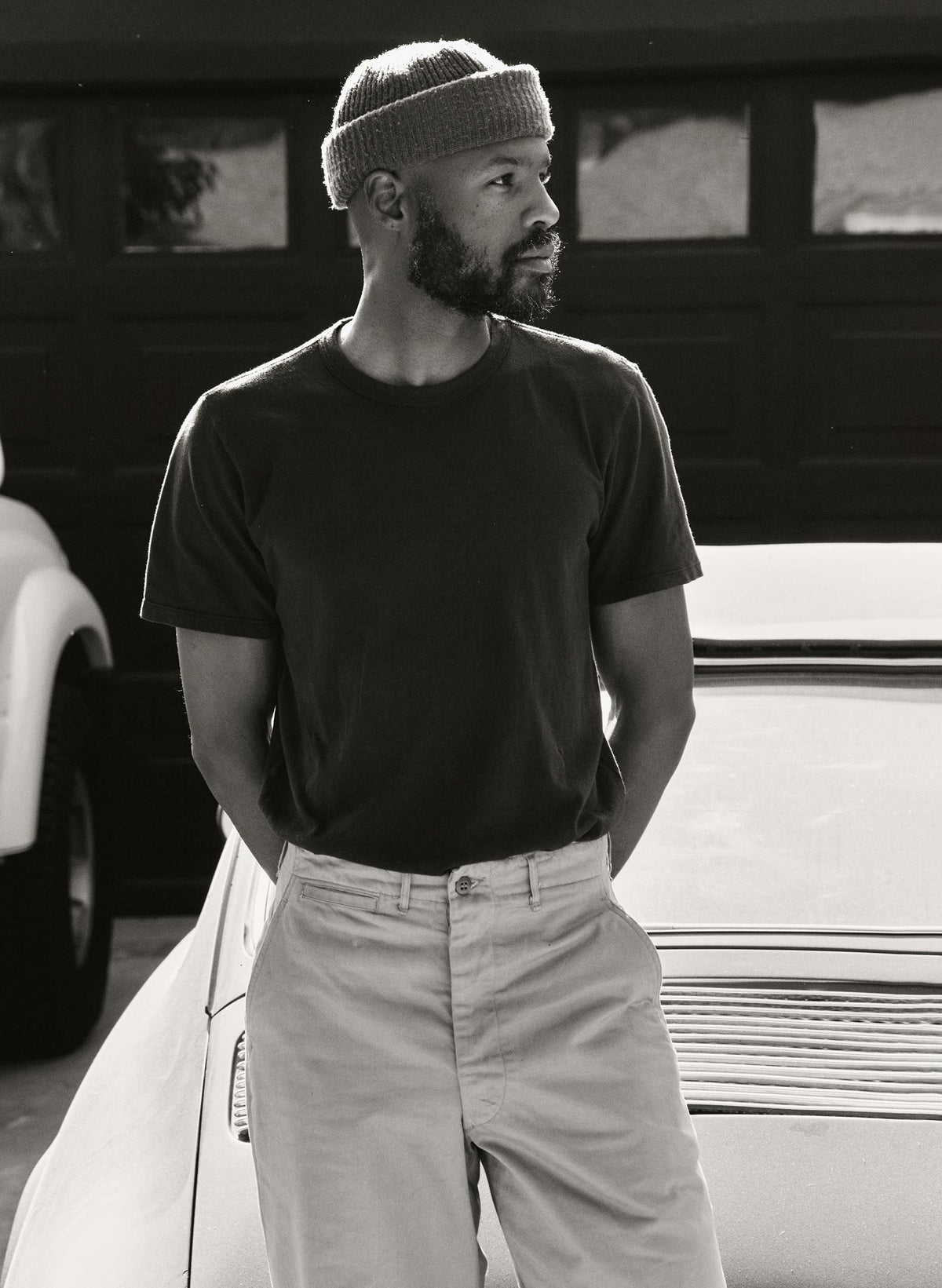 Man wearing the Imogene + Willie Classic Knit Crew in Black, paired with khaki trousers and a knit beanie, leaning against a white vintage car — lifestyle shot, black and white