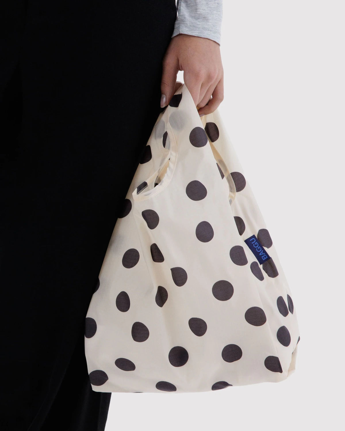 BAGGU Baby Baggu in Cream and Black Polka Dot, held at a model’s side to show the small cream shopping bag with black dots.