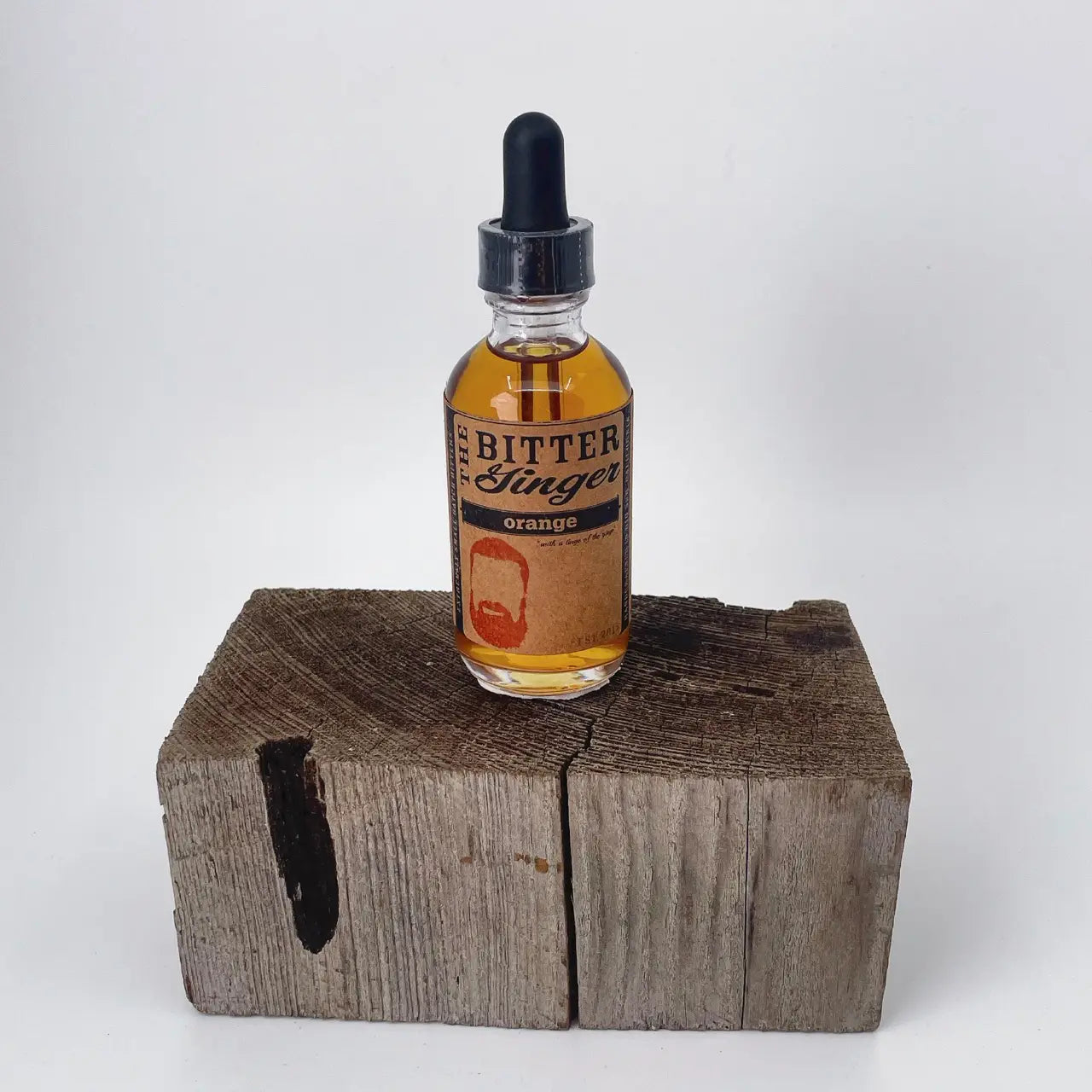 The Bitter Ginger Orange Bitters 2 oz glass dropper bottle with kraft paper label, displayed upright on a rustic weathered wood block against a white background