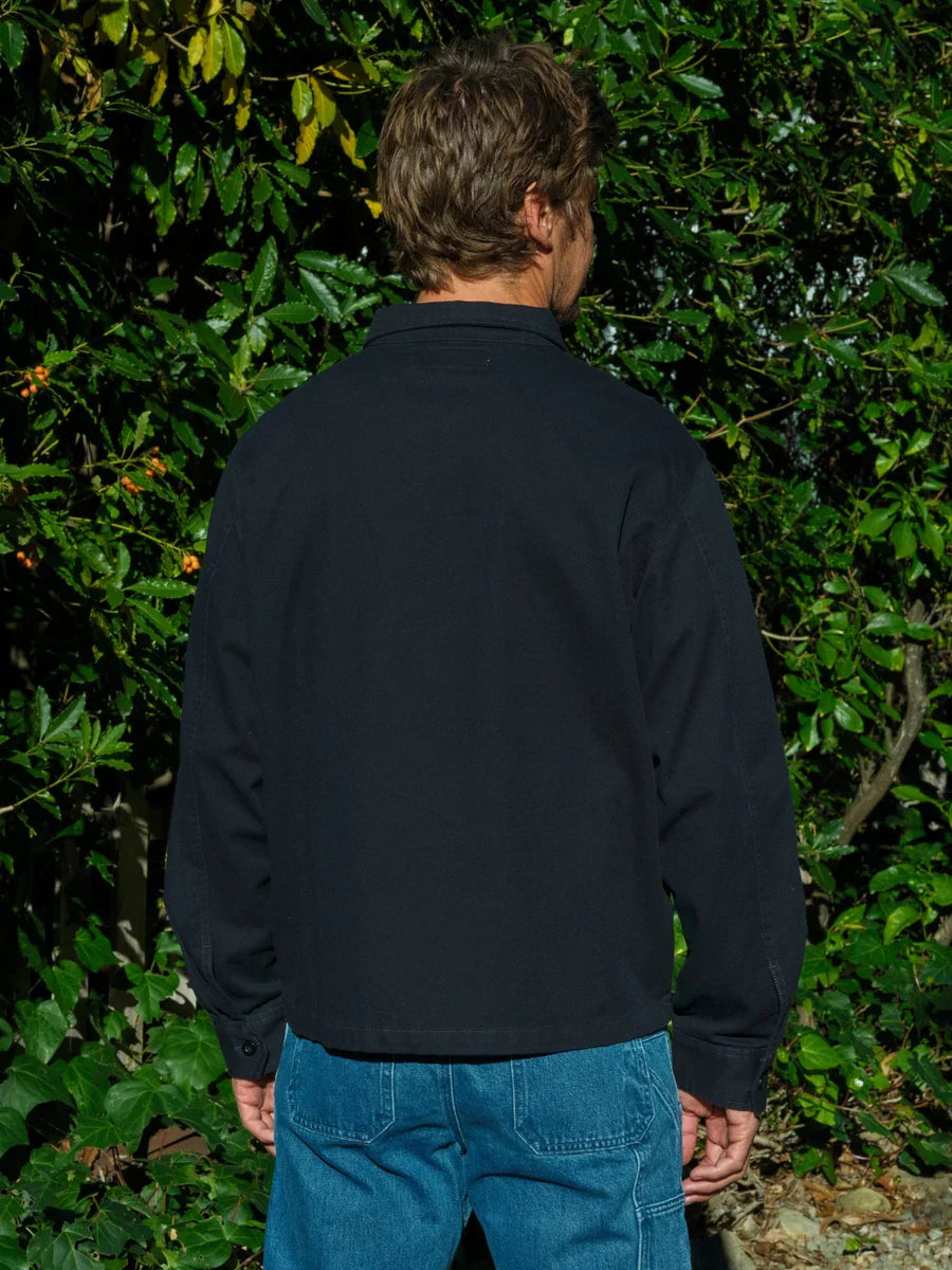 Back view of a man wearing the Mollusk Gas Station Jacket in Navy, showing the clean back panel, spread collar, button cuffs, and relaxed fit in navy 100% cotton twill, styled with bright blue jeans against a garden hedge with red berries