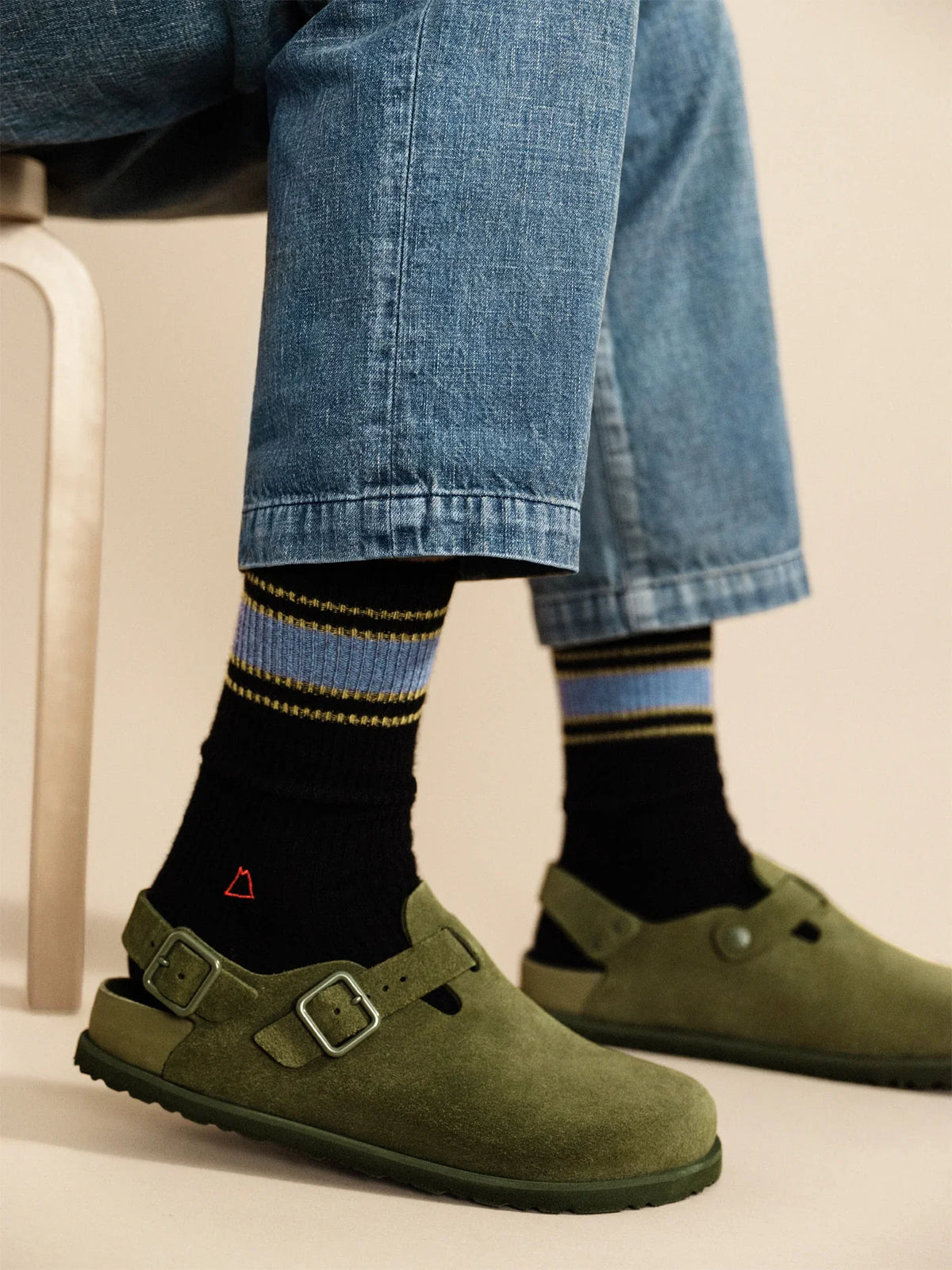Model wearing the Hansel from Basel Athleisure Rib Cashmere Men's Crew in Black, seated lifestyle shot, black ribbed cashmere crew socks with blue and gold stripe cuff and red triangle logo, styled with blue cuffed denim and olive green suede buckle sandals