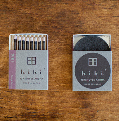 Two Hibi Ylang Ylang Incense Match boxes in soft lavender and grey on a wood surface — one open showing 8 match-style incense sticks with white tips and 'Ylang Ylang' on the spine, one closed with the Hibi 10 Minutes Aroma logo, Made in Japan