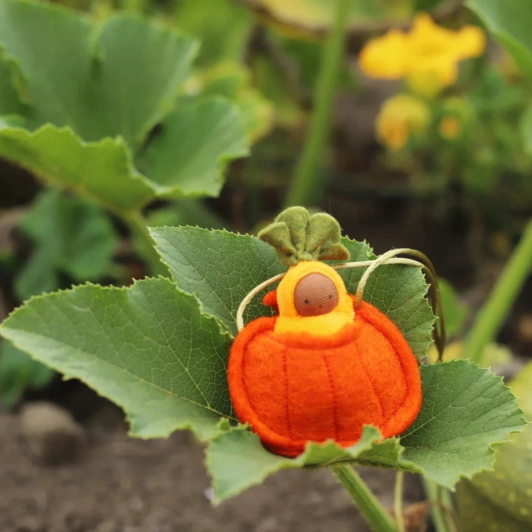 Fairy Shadow Pumpkin Seedling Necklace in tan skin tone, resting on a green pumpkin leaf in a garden, showing a knitted tan-skin seedling doll in a yellow hood cradled in a felted orange ribbed pumpkin pouch with green felt leaf top and natural cord strap